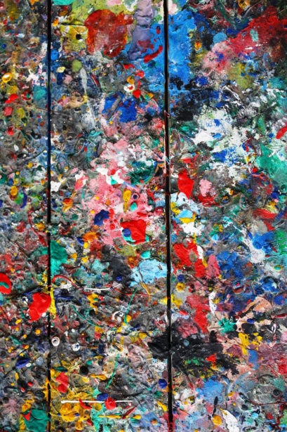 Abstract art with vibrant splashes of red, blue, yellow, and green paint on weathered wood panels, suggesting a messy artist's workspace.