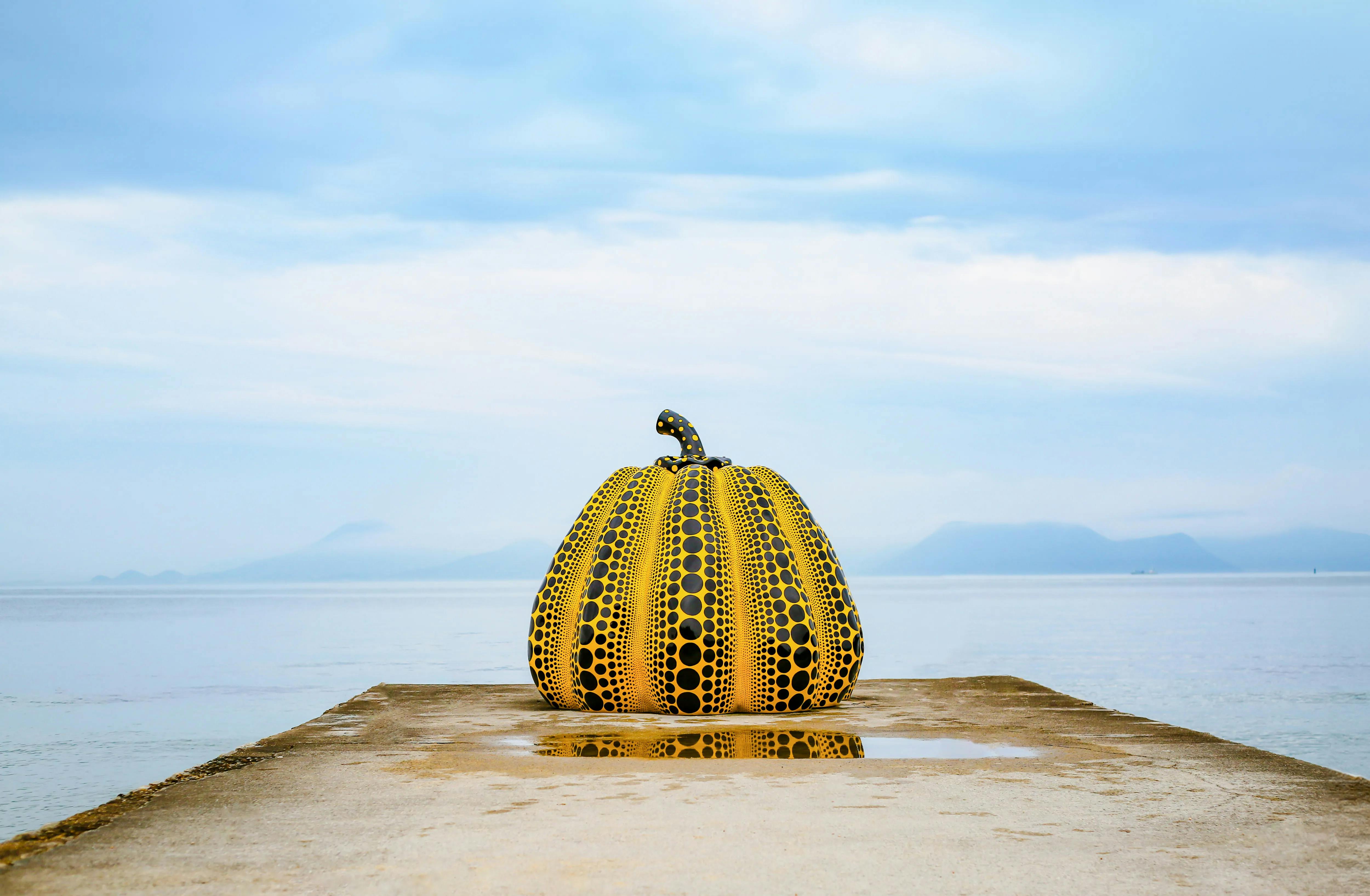 Yellow pumpkin sculpture with black polka dots on a pier in Naoshima, Japan, with the sea and sky in the background.