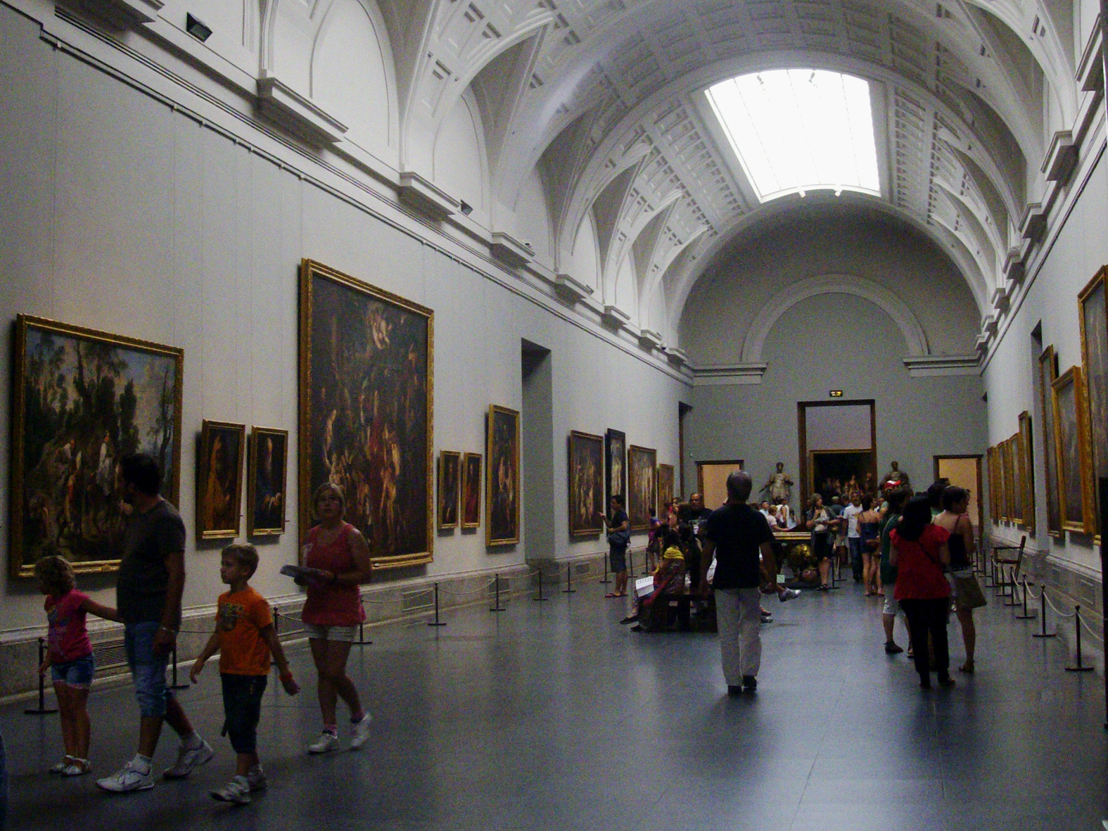 Interior view of the Prado Museum's permanent collection gallery with visitors viewing large, framed paintings under a high, arched ceiling with a skylight.