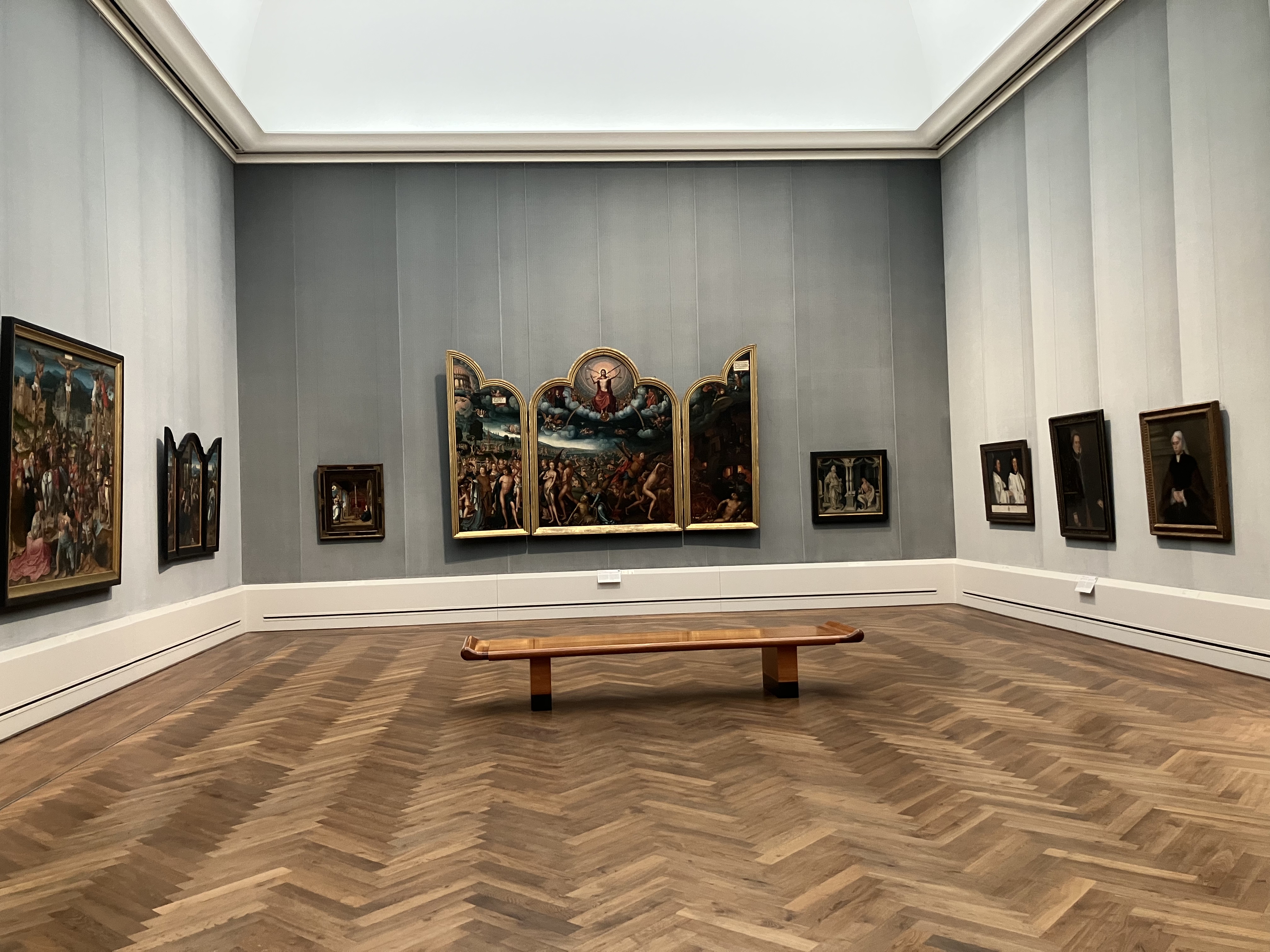 Interior view of Room VI at the Gemäldegalerie in Berlin, showcasing a collection of Old Master paintings, including a large triptych and several portraits, displayed on a grey wall with a wooden parquet floor and a central bench.