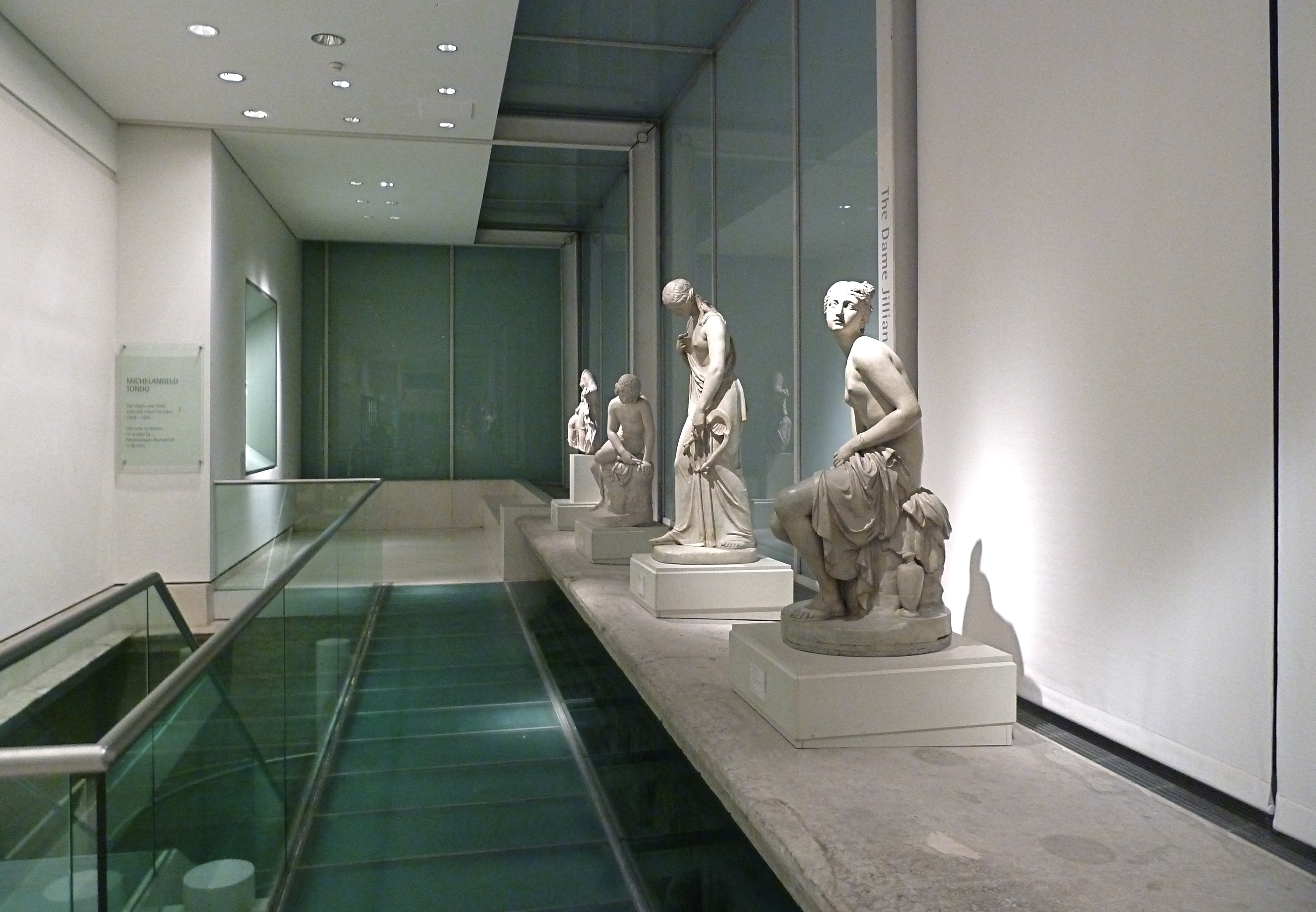 Interior view of the Royal Academy sculpture gallery, showcasing classical marble statues displayed on a raised platform with a glass floor below.