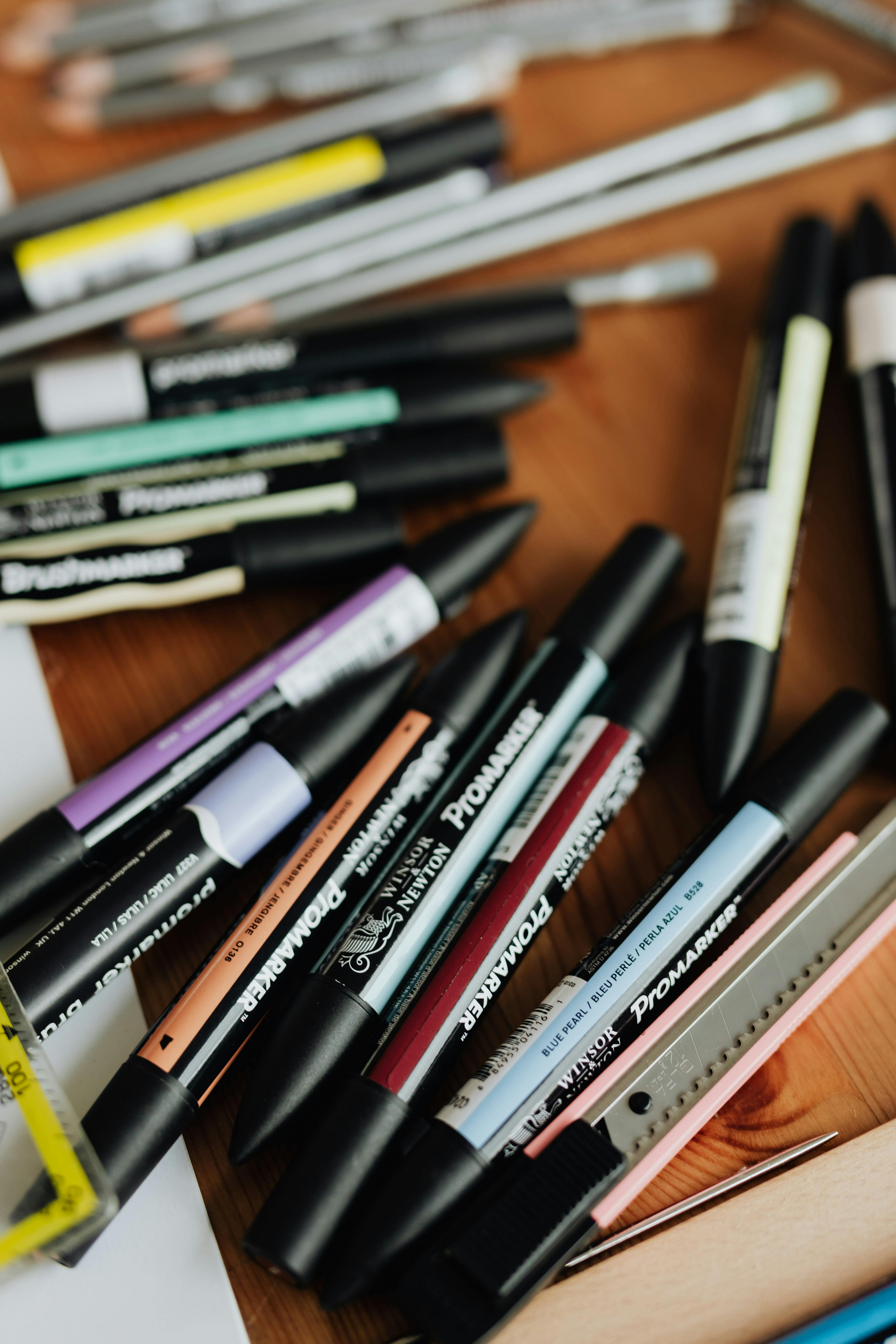 A collection of Winsor & Newton Promarker alcohol-based markers in various colors, along with pencils and a craft knife, arranged on a wooden surface, ideal for artists.