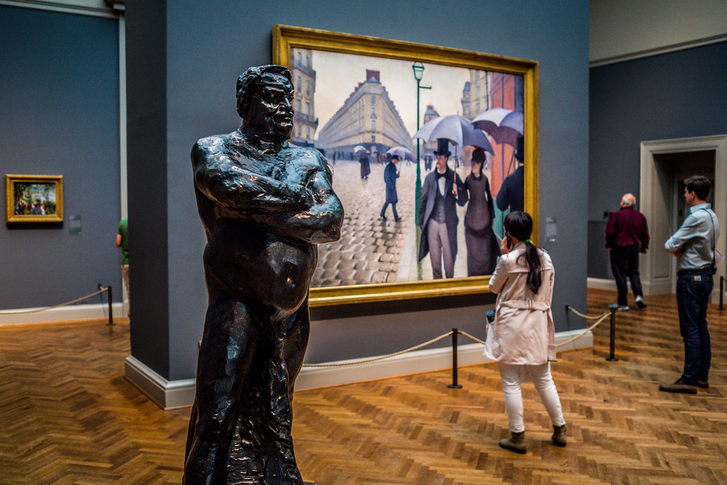 A bronze statue of a man with his arms crossed stands in the foreground at the Art Institute of Chicago, with a painting of a Parisian street scene and museum visitors in the background.