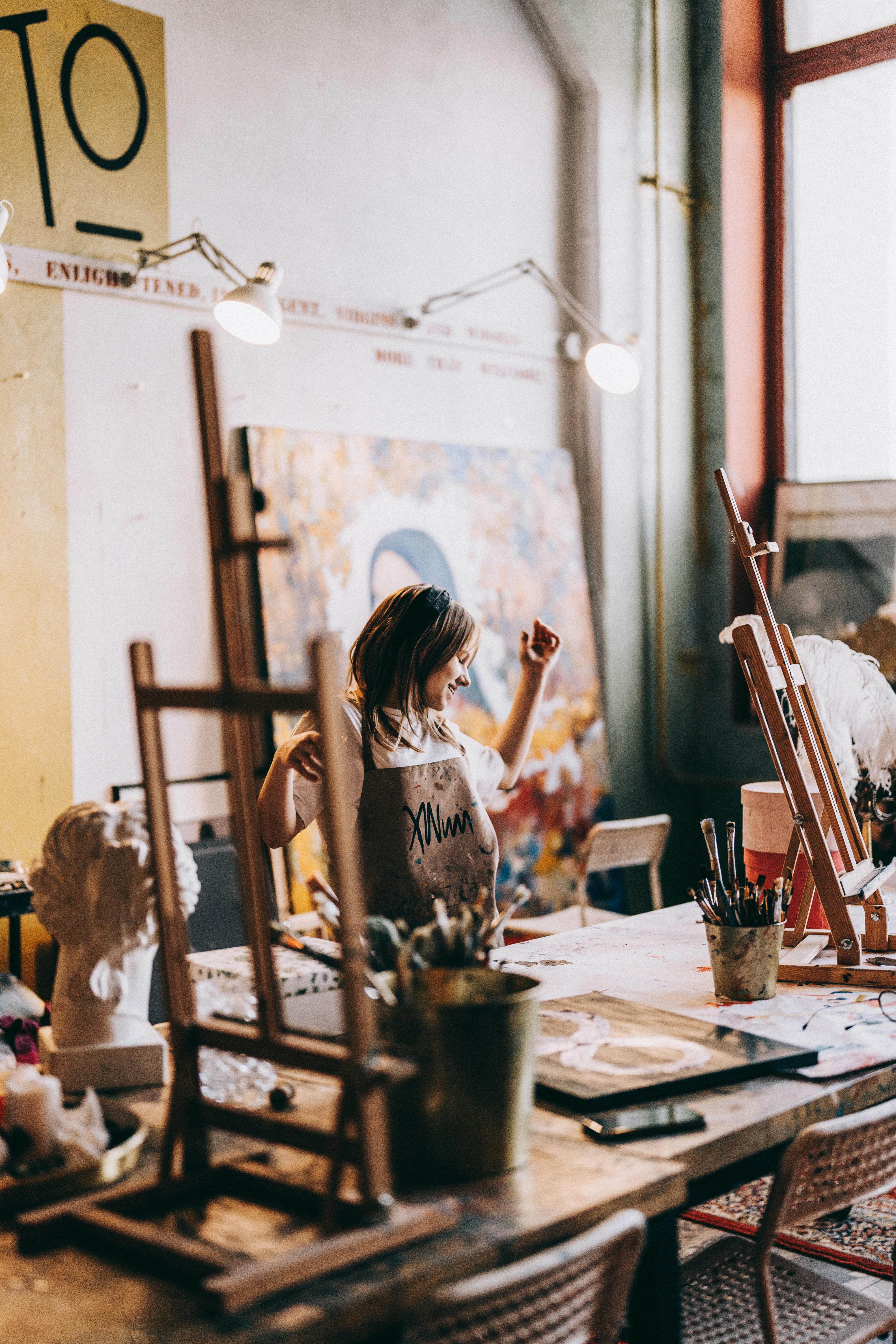 Young woman joyfully painting in a cluttered art studio, surrounded by easels and art supplies.