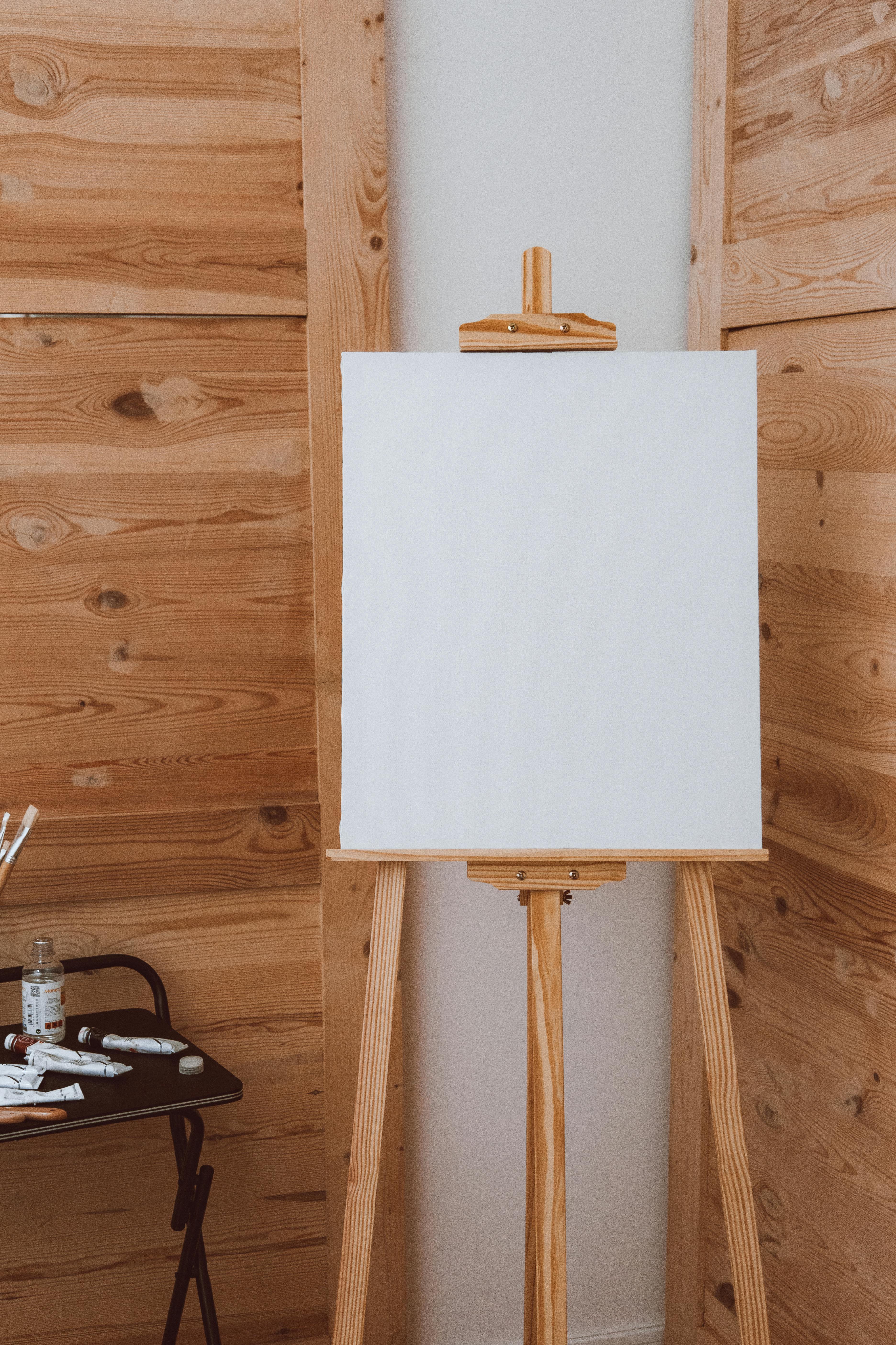 A white canvas sits on a wooden easel, with art supplies like paint tubes and brushes on a nearby table, set against a warm wooden background.