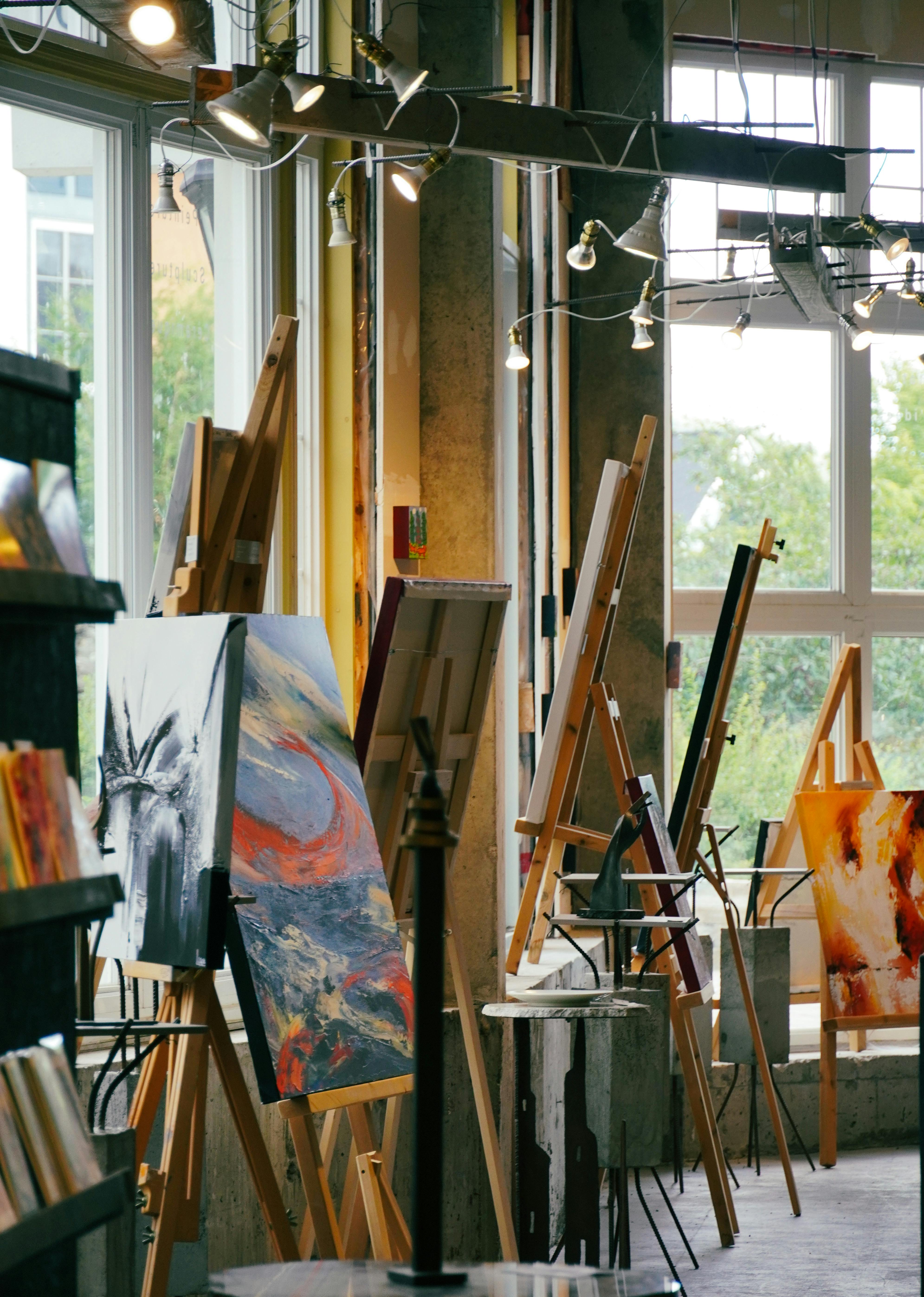 Artists' studio interior with multiple wooden easels displaying vibrant abstract paintings, illuminated by industrial-style studio lights near large windows.