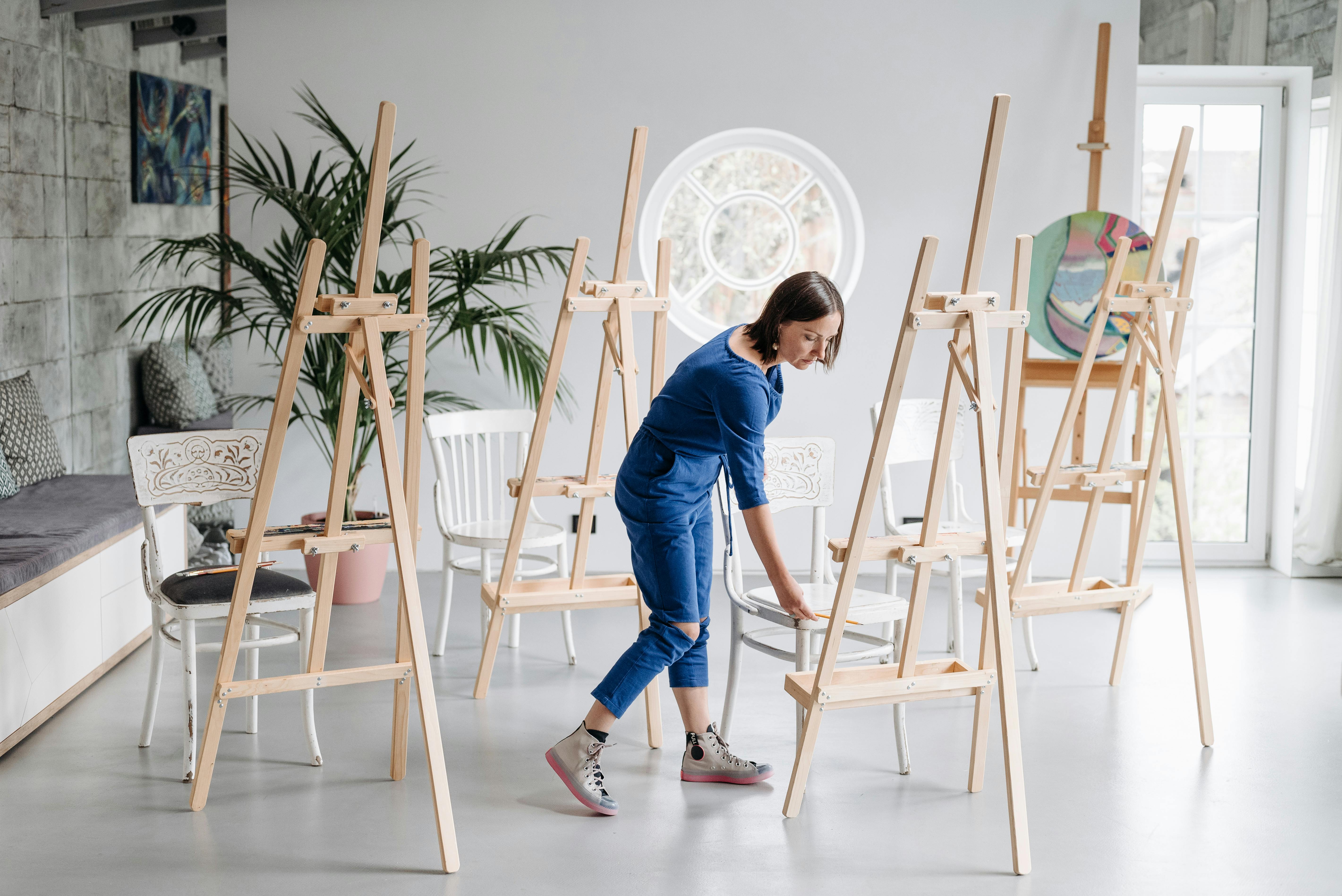 A woman in a blue jumpsuit inspects wooden easels in a bright, spacious art studio with large windows and plants.