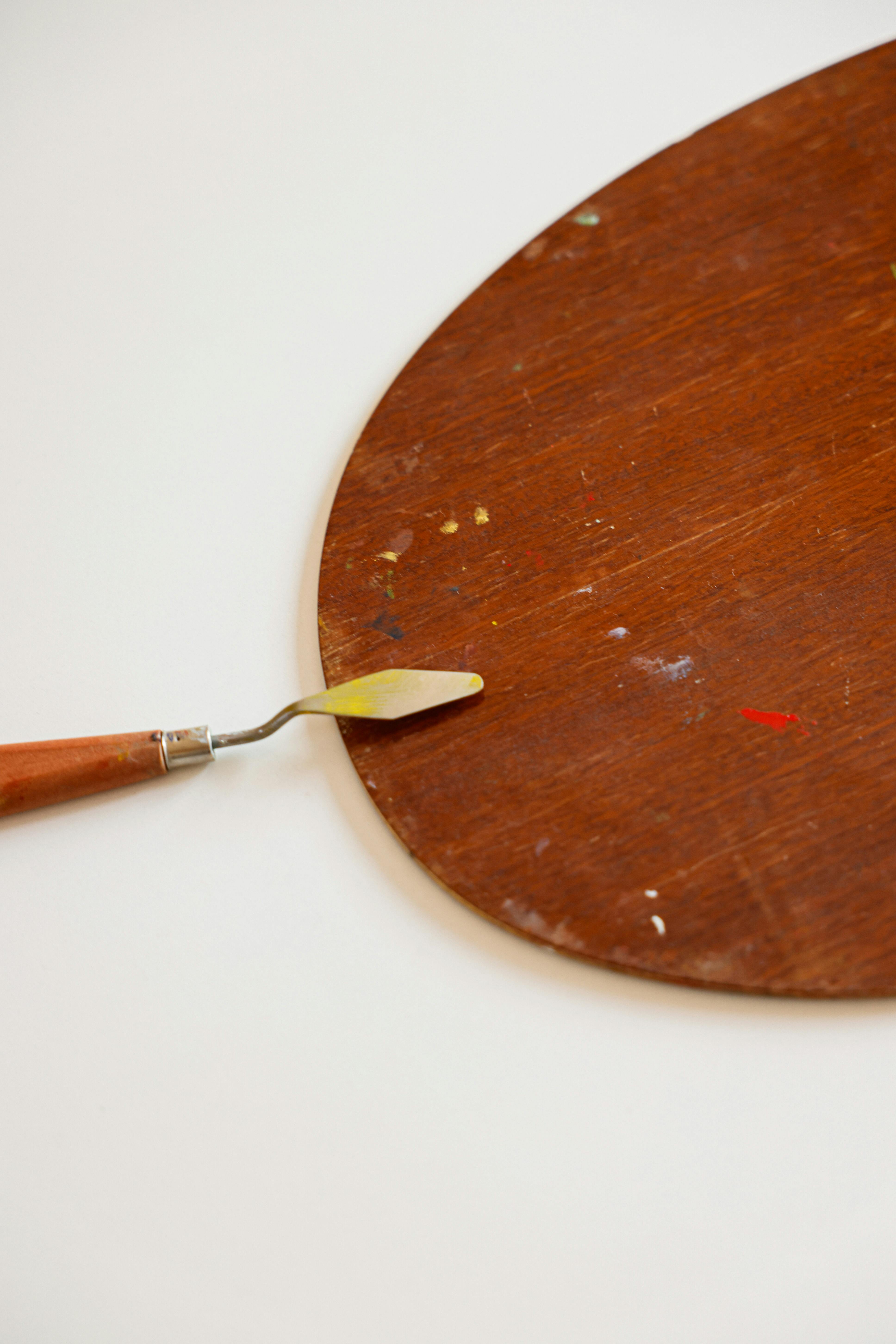 A palette knife with a yellow tip rests on a wooden artist's color mixing palette, which has small specks of paint on its surface.
