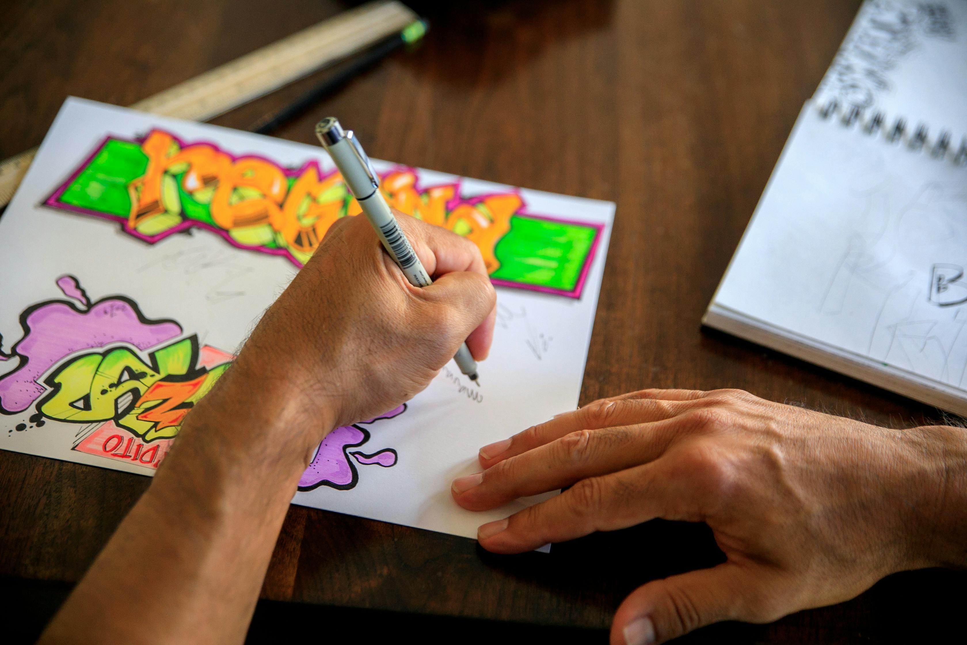 Graffiti artist drawing with colorful markers on paper, with a sketchbook in the background.