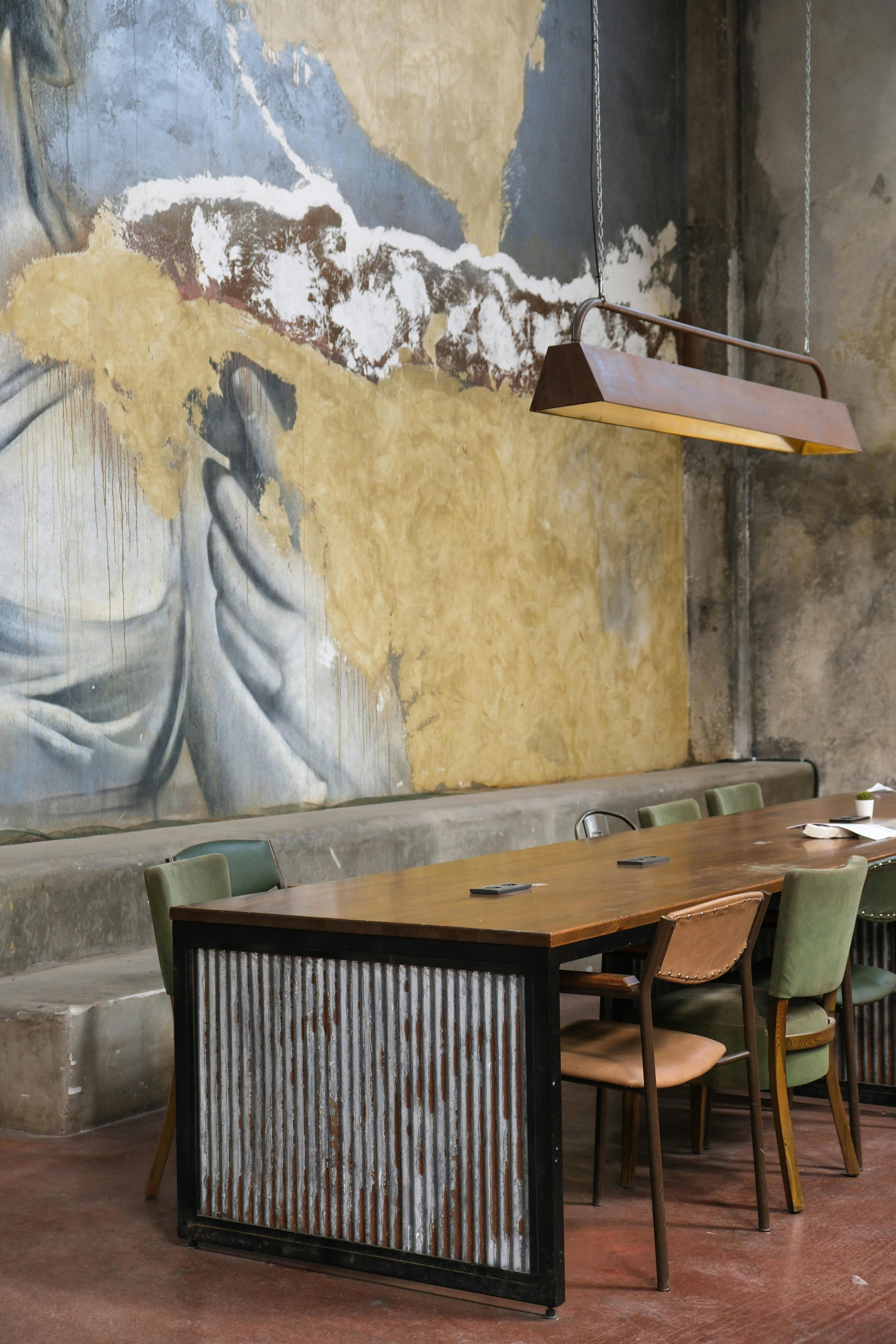 Interior Design with Art on Wall: Filled with color and creativity, this mural transforms a weathered wall in a public setting. Decorating large walls with art, the scene combines bold strokes of white and blue against a backdrop of textured, aged brickwork. The dynamic interplay of light on the mural highlights its artistic flair, making it a focal point in the room’s industrial-chic ambiance. The weathered table and vibrant green chairs below complement the painting, creating a harmonious blend of urban art and functional design.