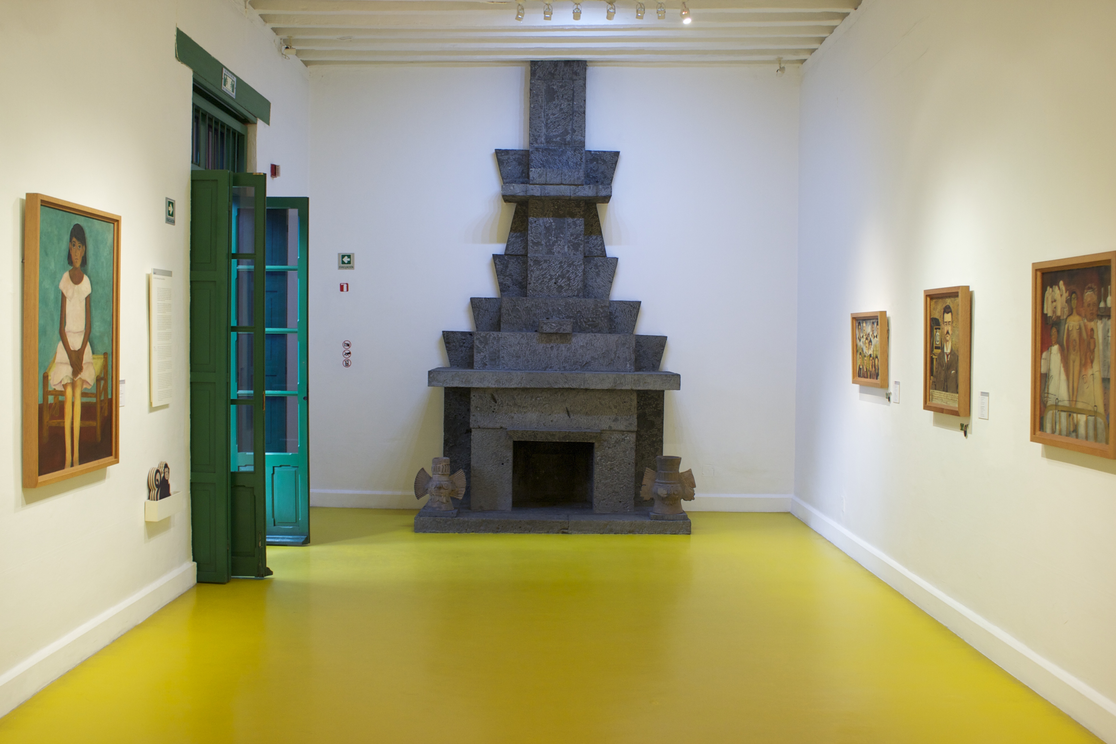 Interior of Room 1 at the Frida Kahlo Museum (Casa Azul) in Coyoacan, Mexico City, featuring a large stone fireplace, paintings on the wall, and a bright yellow floor.
