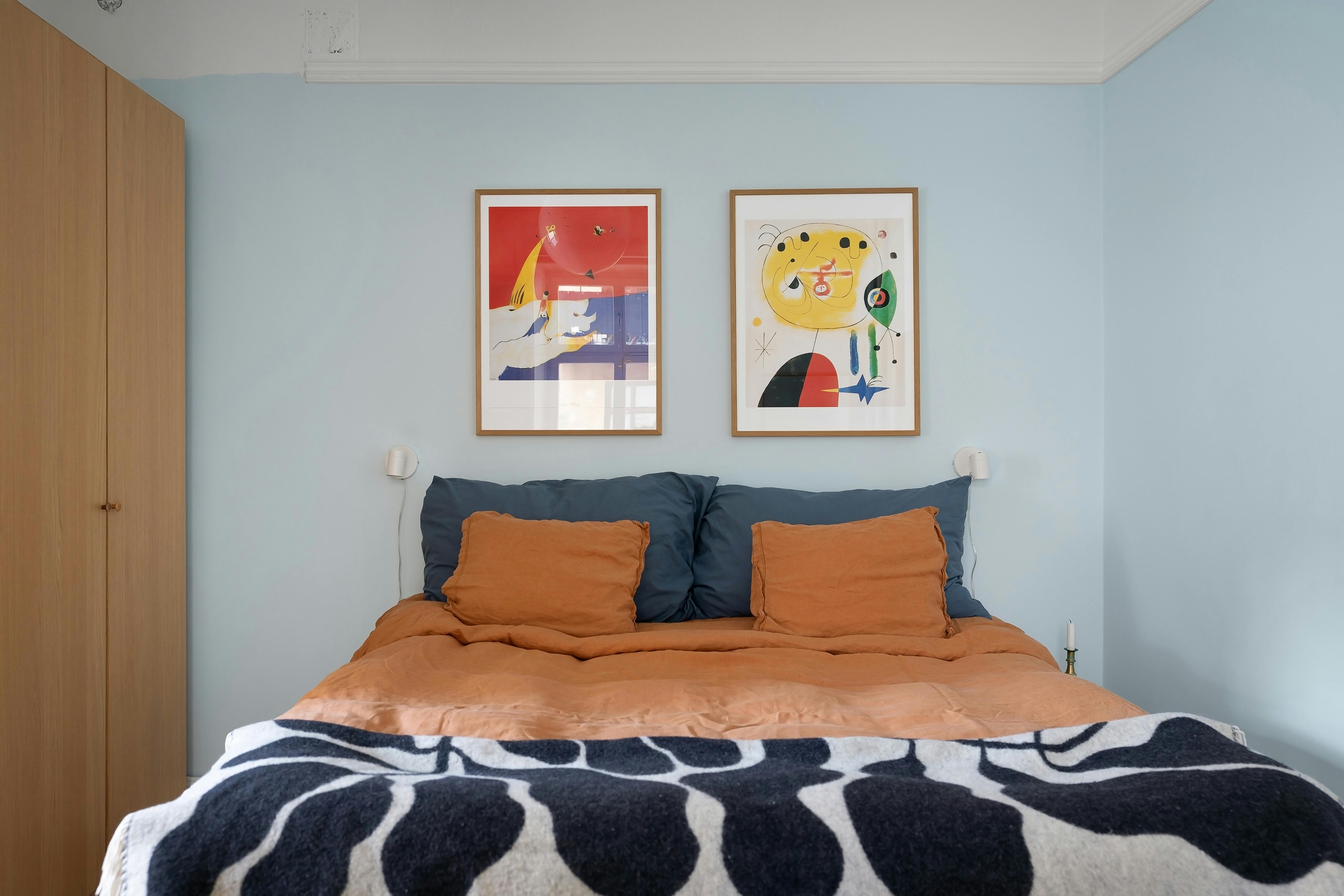 Two framed abstract art pieces hanging on a light blue wall above a bed with orange and blue bedding and a black and white patterned blanket.