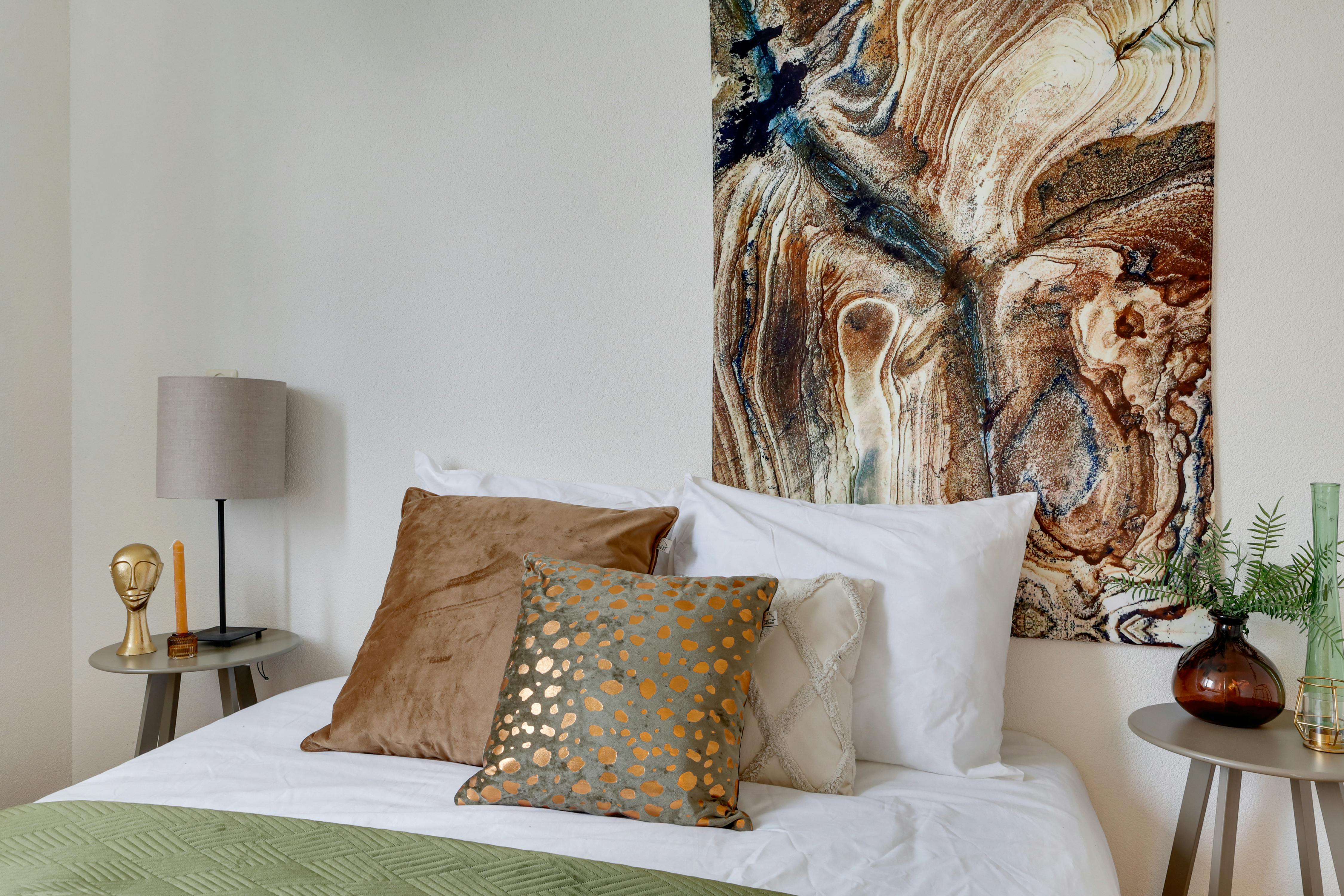 Bedroom with a large abstract painting above the bed, featuring warm earth tones and blue accents. The bed is made with white linens and several decorative pillows, including a brown velvet pillow and a grey pillow with gold spots. A green quilt is folded at the foot of the bed. Beside the bed are nightstands with a lamp, a candle, and a vase with greenery.