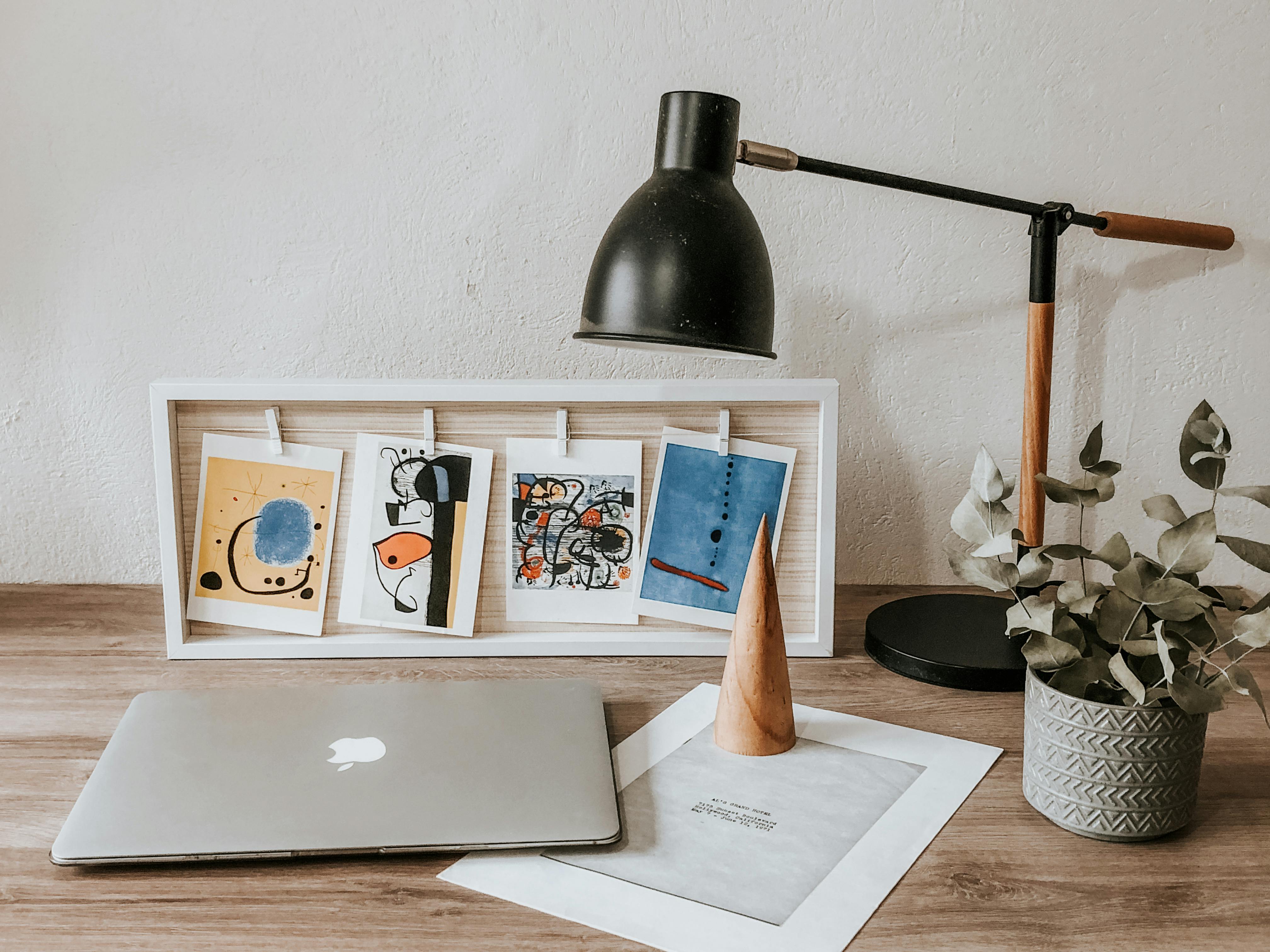 Modern home office desk setup with a laptop, abstract art prints in a white frame, a black desk lamp, and a potted eucalyptus plant.