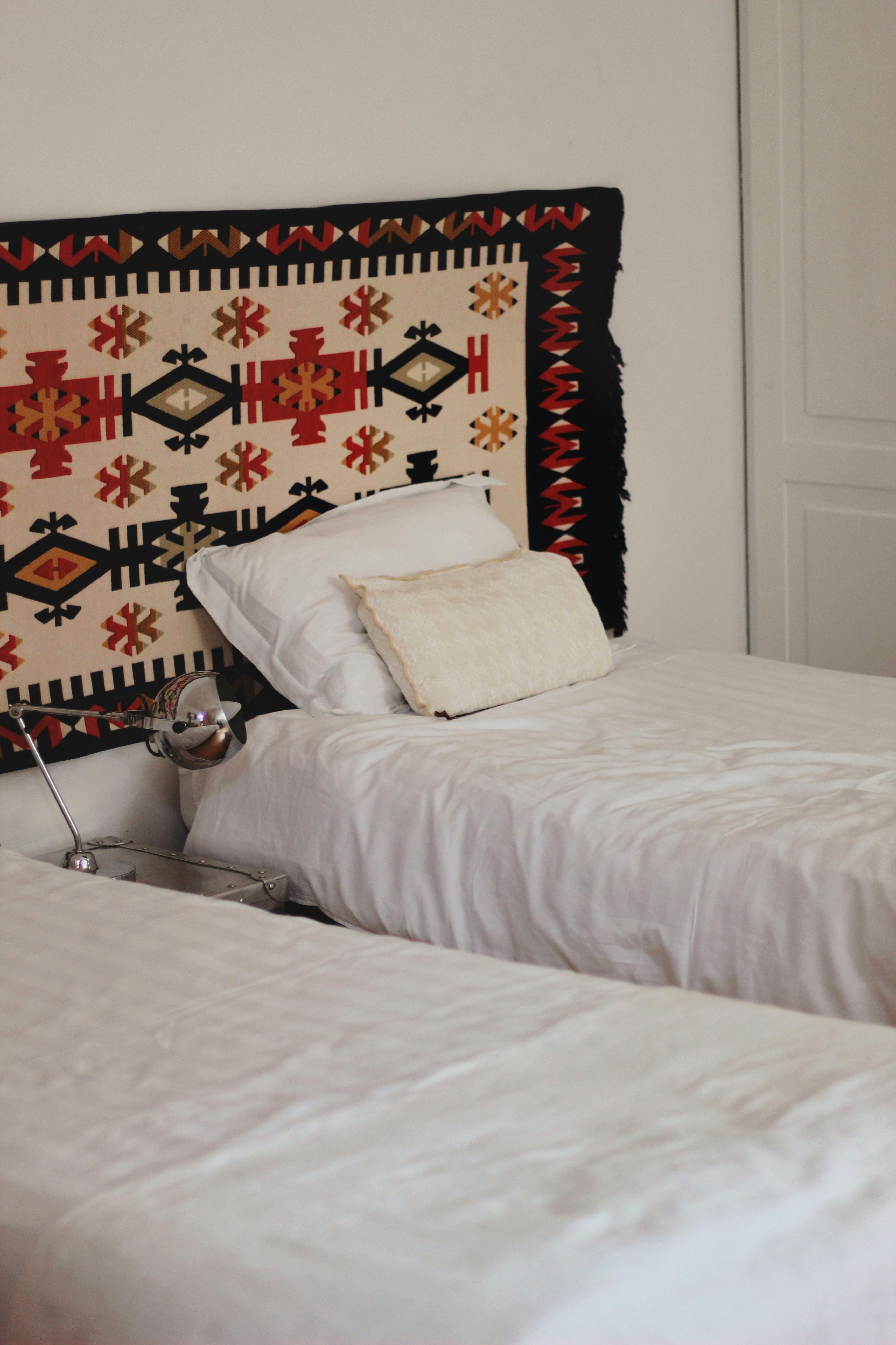 Hotel room interior featuring two twin beds with white bedding, a decorative tapestry above the headboards, and a chrome table lamp.