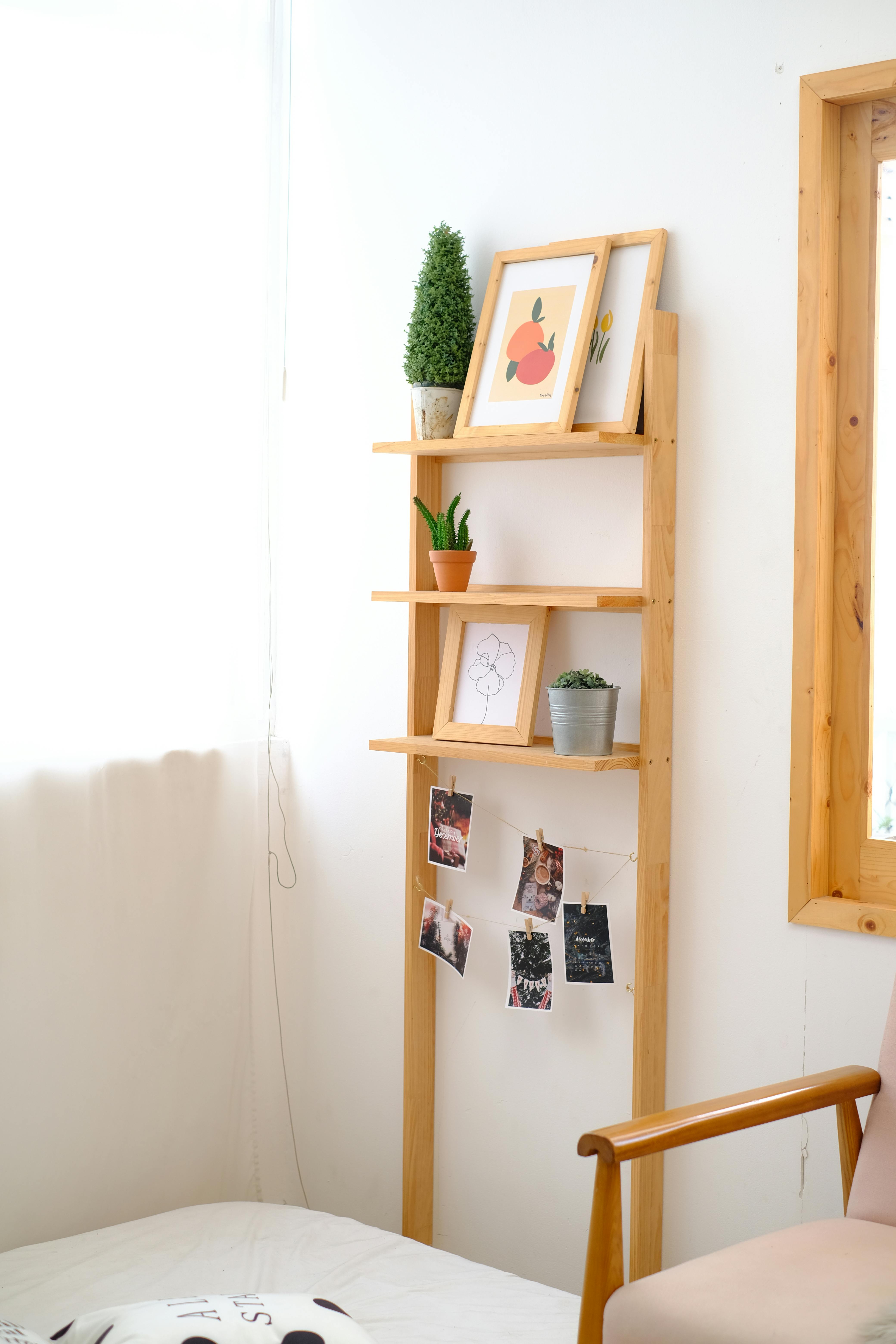 Brown wooden ladder shelf displaying framed art, potted plants, and photos clipped to a string.