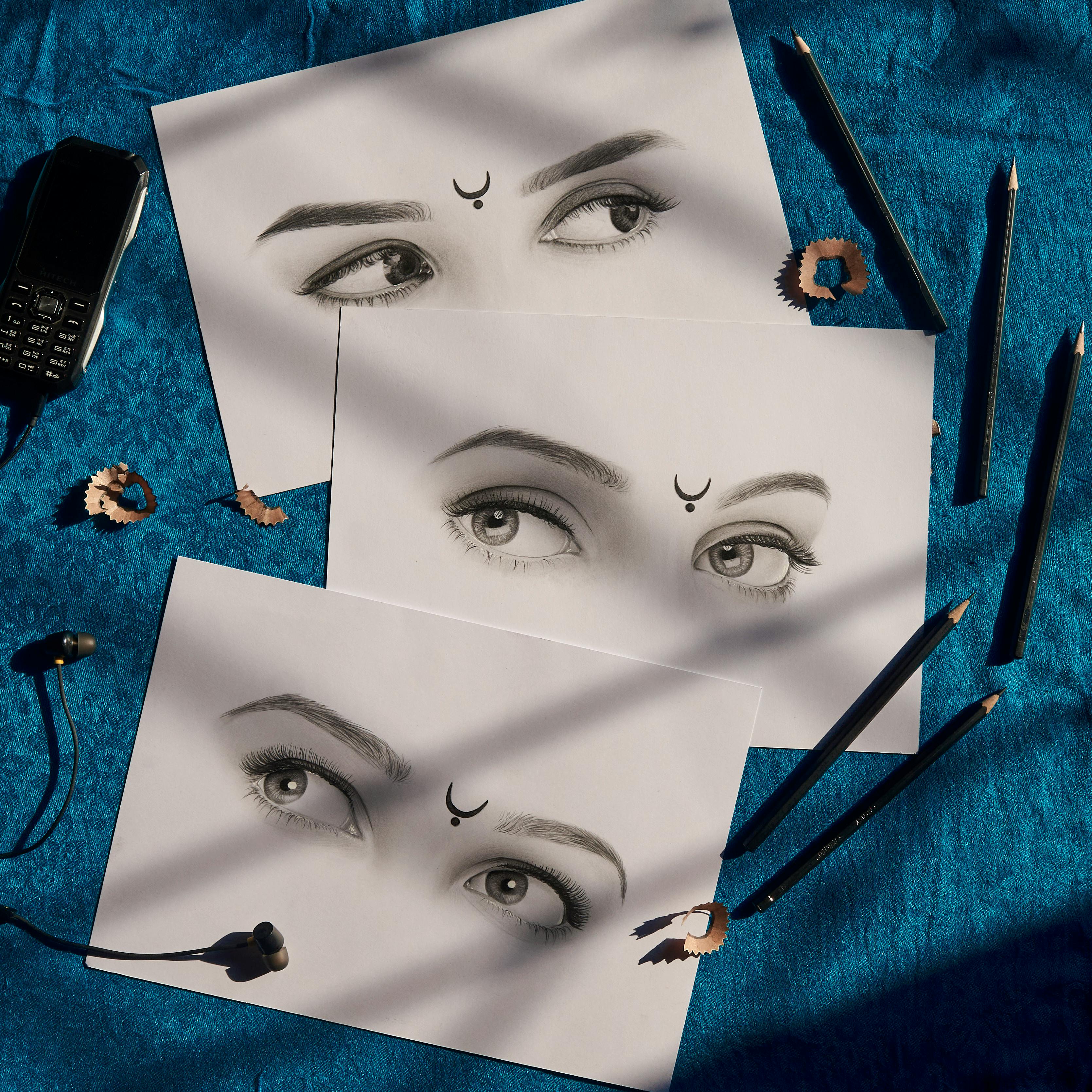 Realistic eye drawing tutorial supplies: pencils, sharpener shavings, earphones, and an old mobile phone on a blue textured surface.