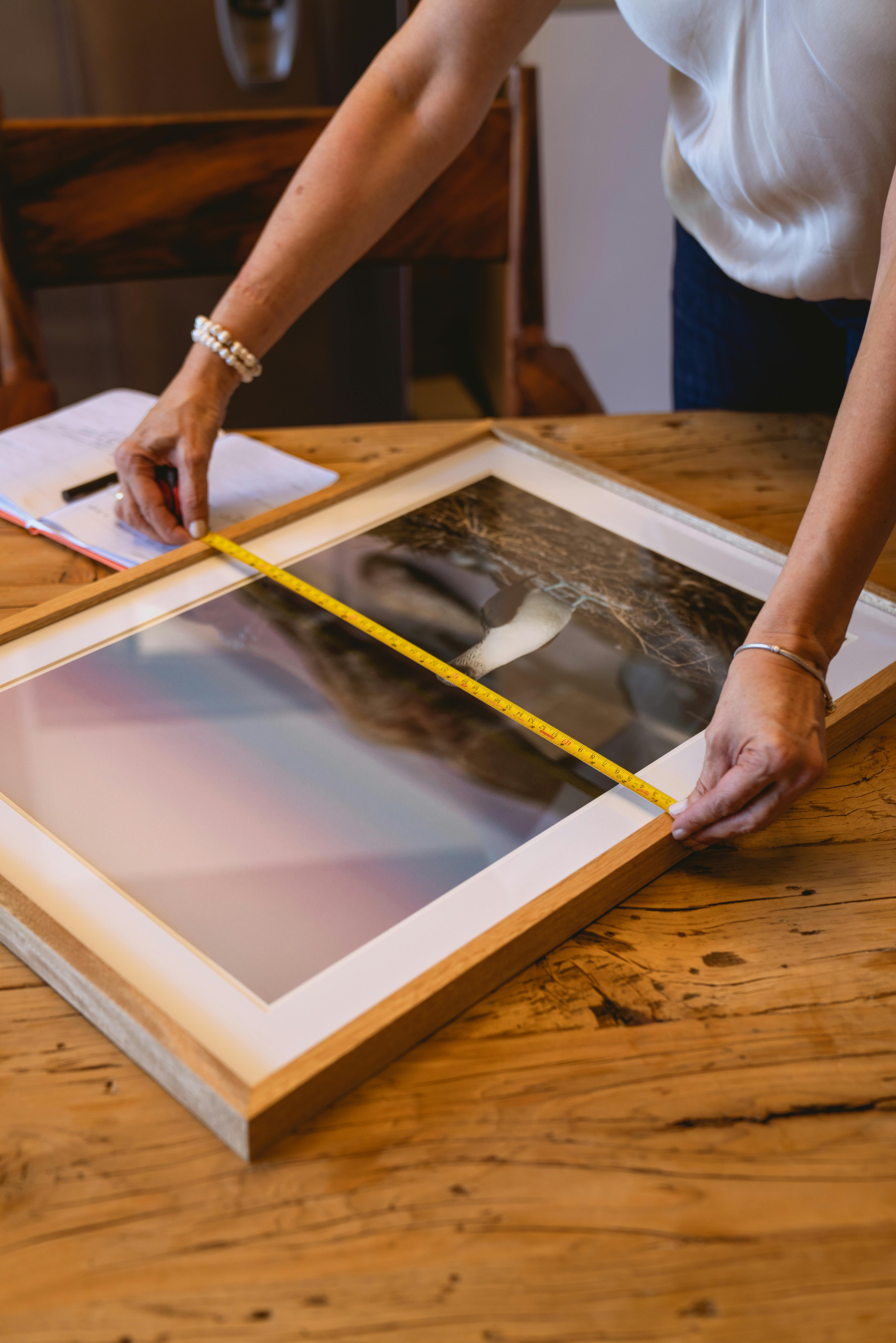 Person measuring a picture frame with a tape measure on a wooden table.