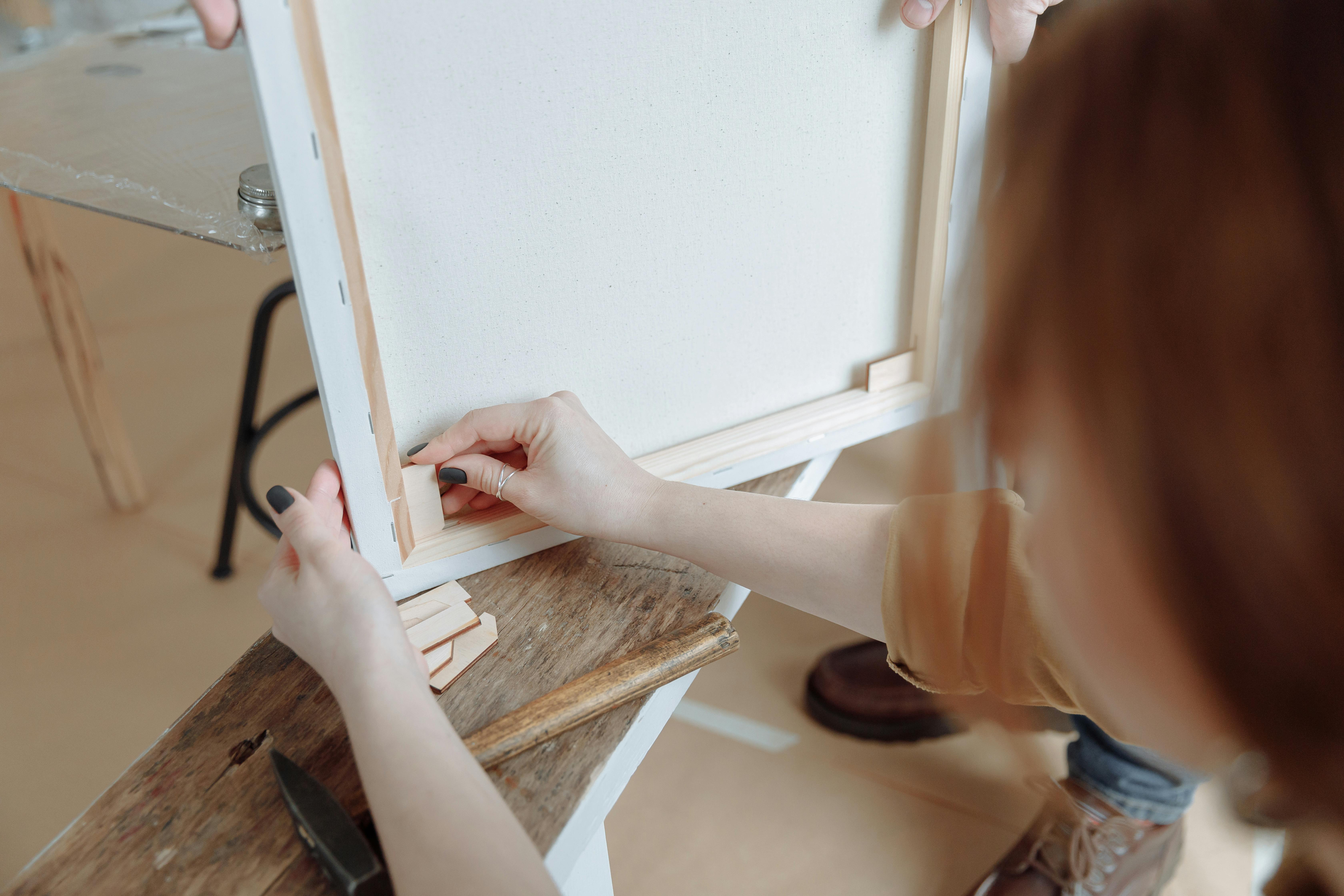 A woman's hands carefully stretching a white canvas onto a wooden frame, preparing it for painting.