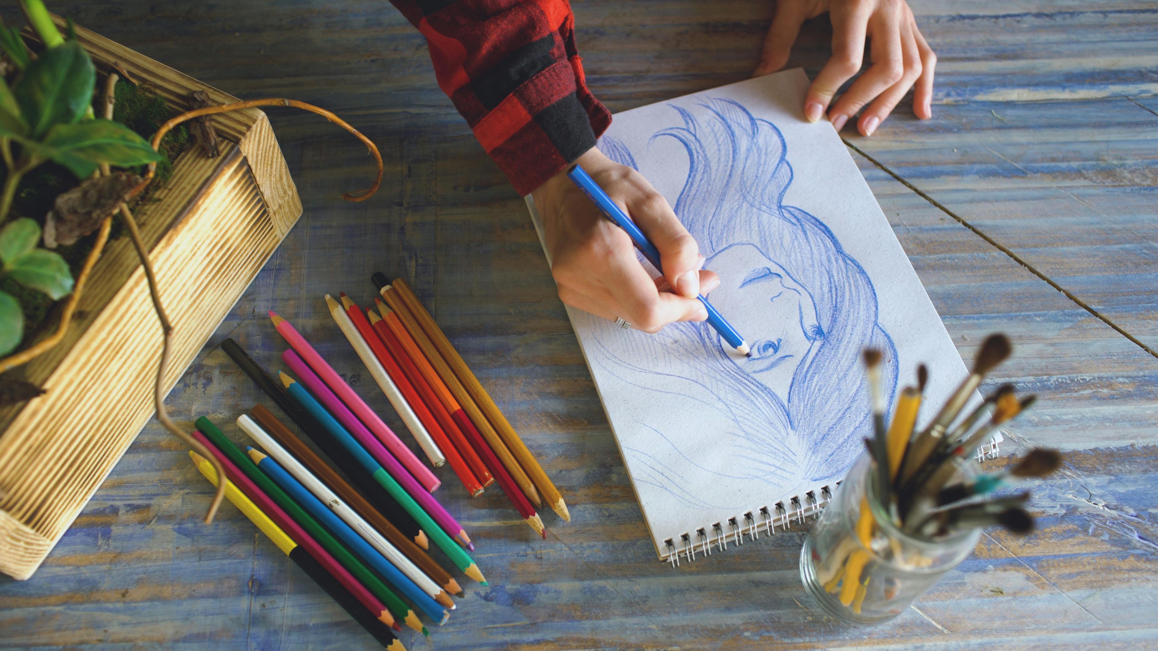 Artist's hands sketching with colored pencils in a sketchbook on a wooden table, capturing a moment or idea