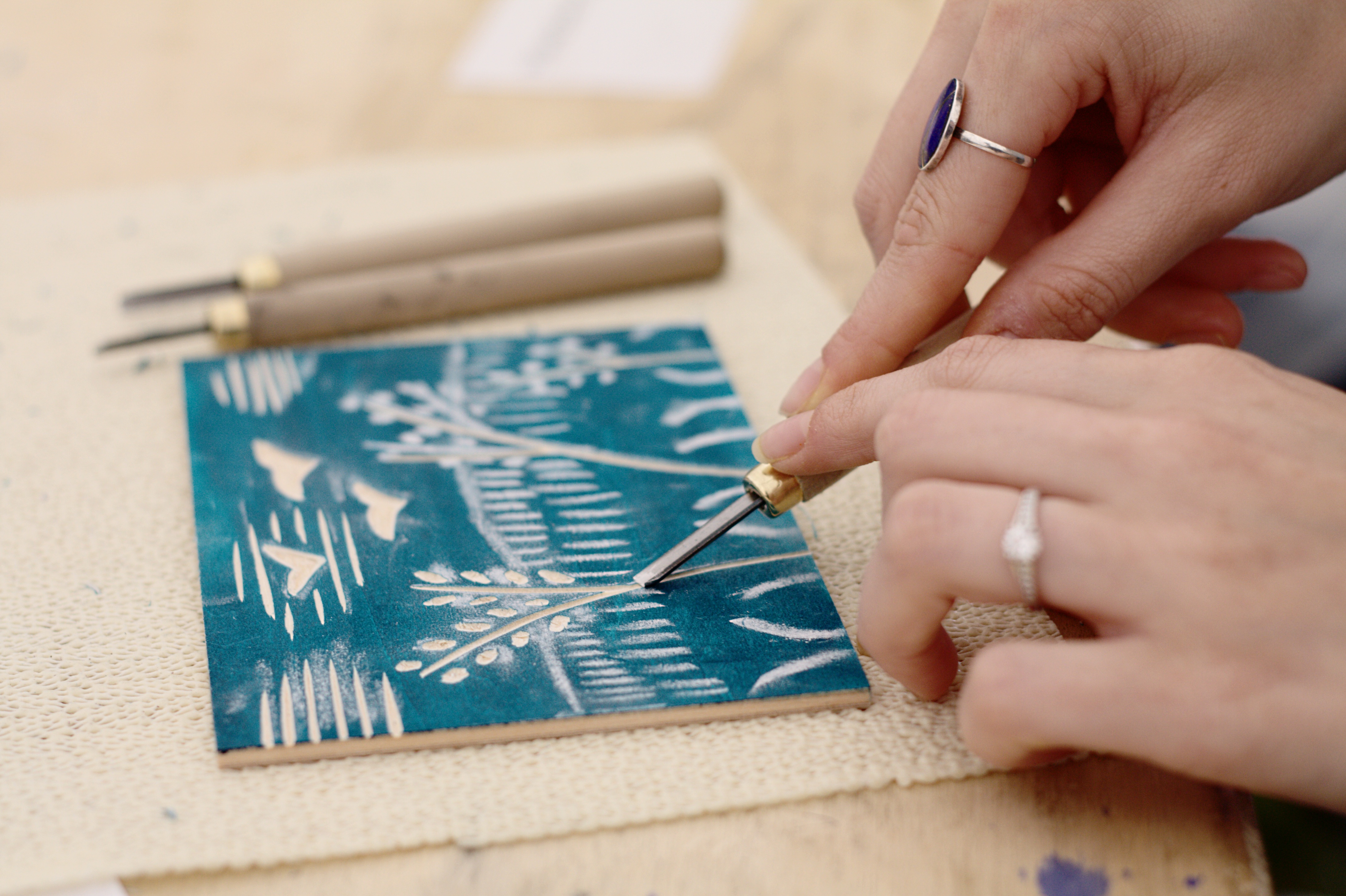 Close-up of hands carving a woodblock print with a tool, showing intricate details of the design and the carving process.