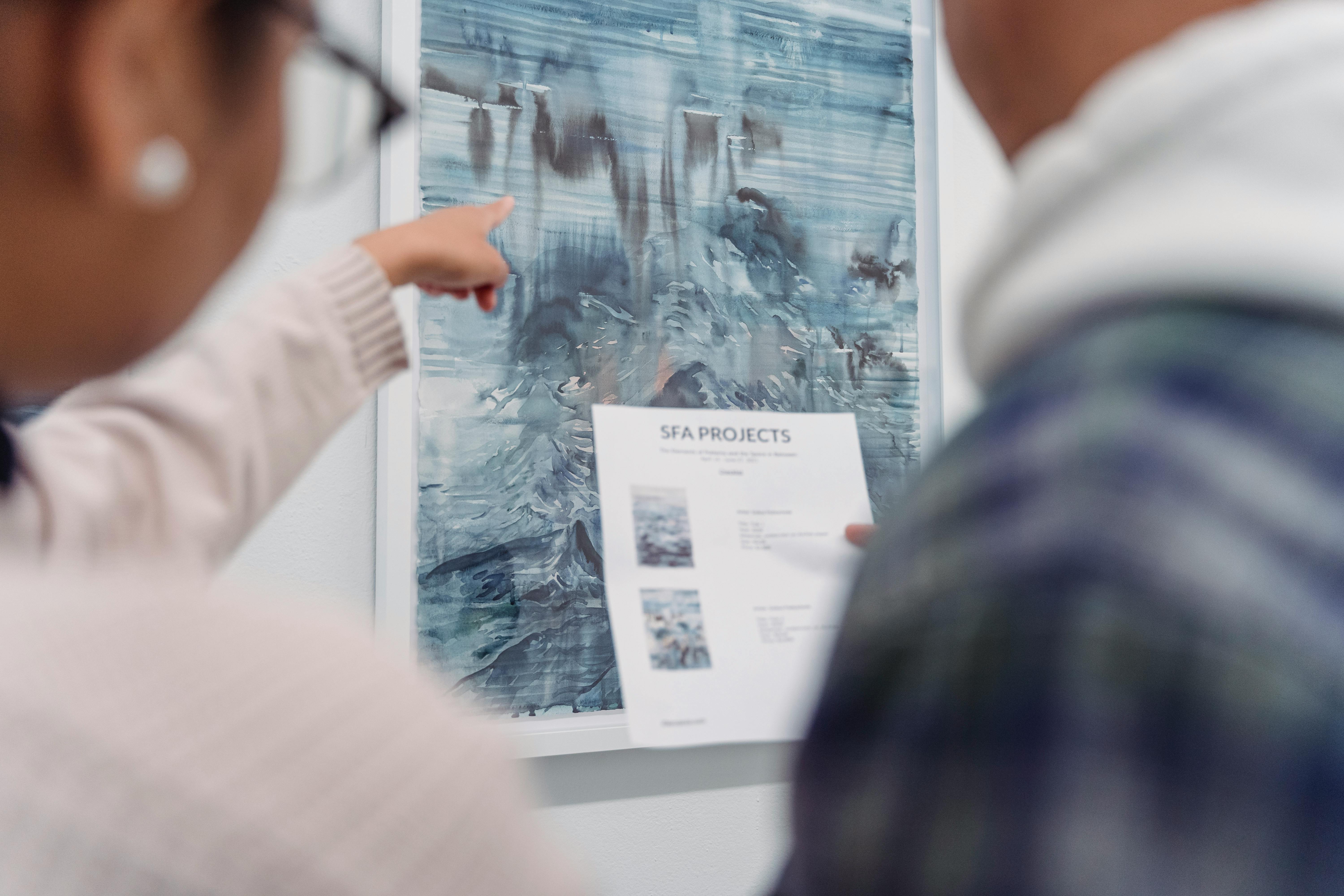 A person pointing their finger at a blue and grey abstract painting in an art gallery.