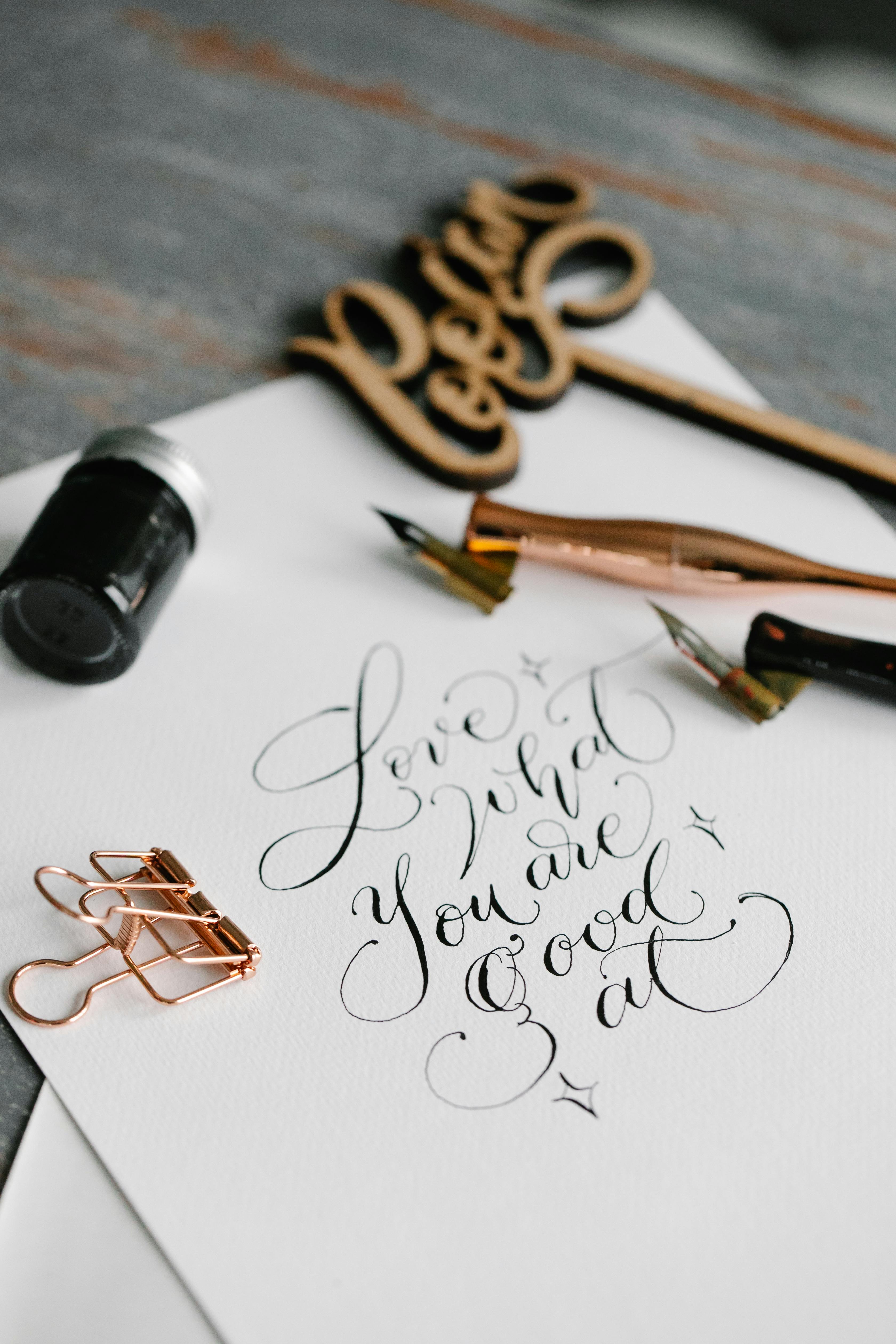 Close-up of calligraphy tools including ink, nibs, and a wooden flourish, with the handwritten quote 'Love what you are good at' on textured white paper.