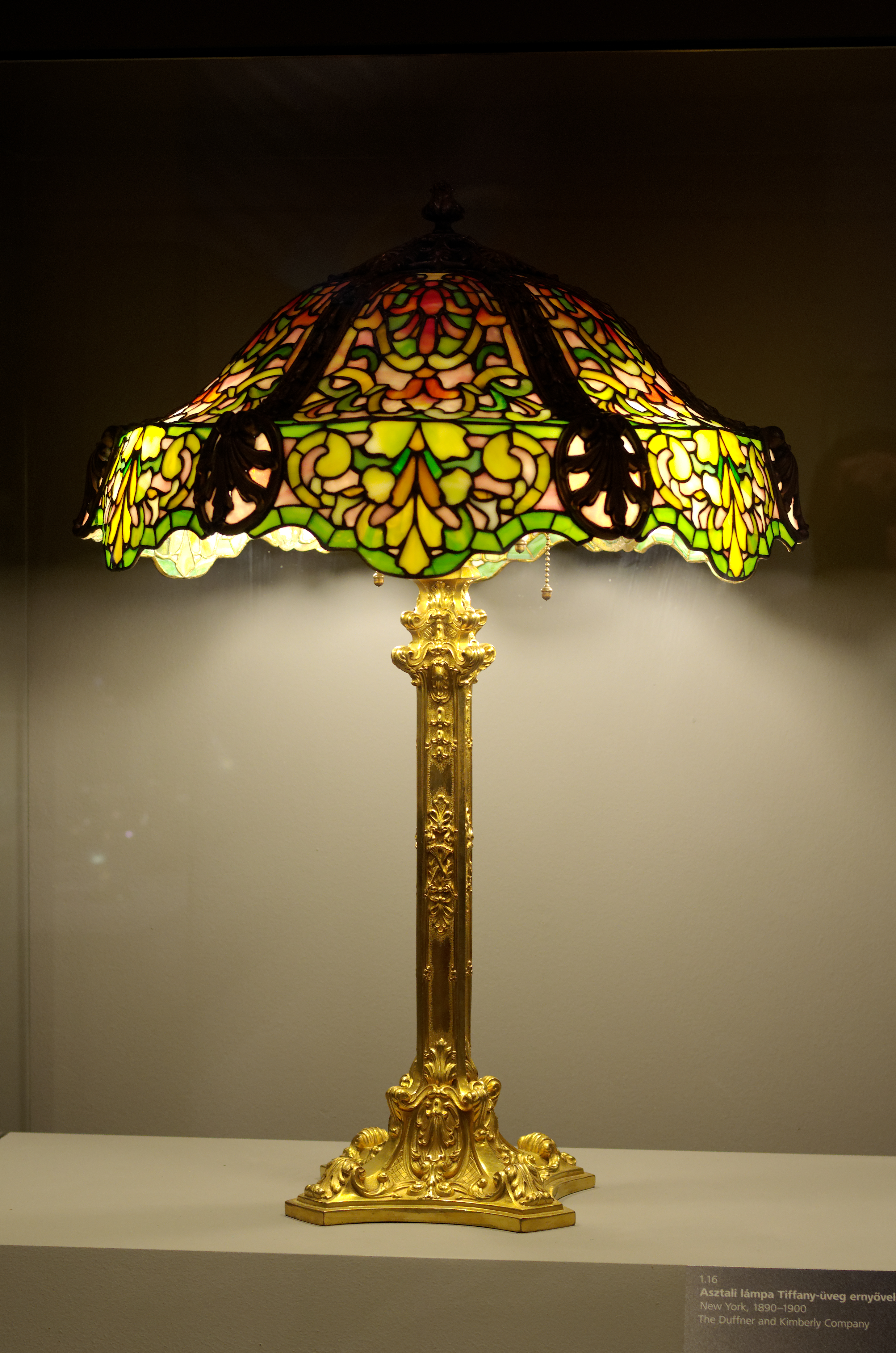 A Tiffany stained glass lamp with an ornate golden base, showcasing intricate floral patterns in green, yellow, and red glass.