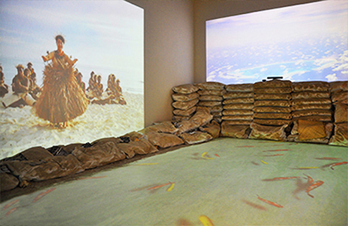 Interactive installation at the 57th Venice Biennale featuring projected images of a traditional dance and clouds, with sandbags forming a barrier and koi fish projected on the floor.