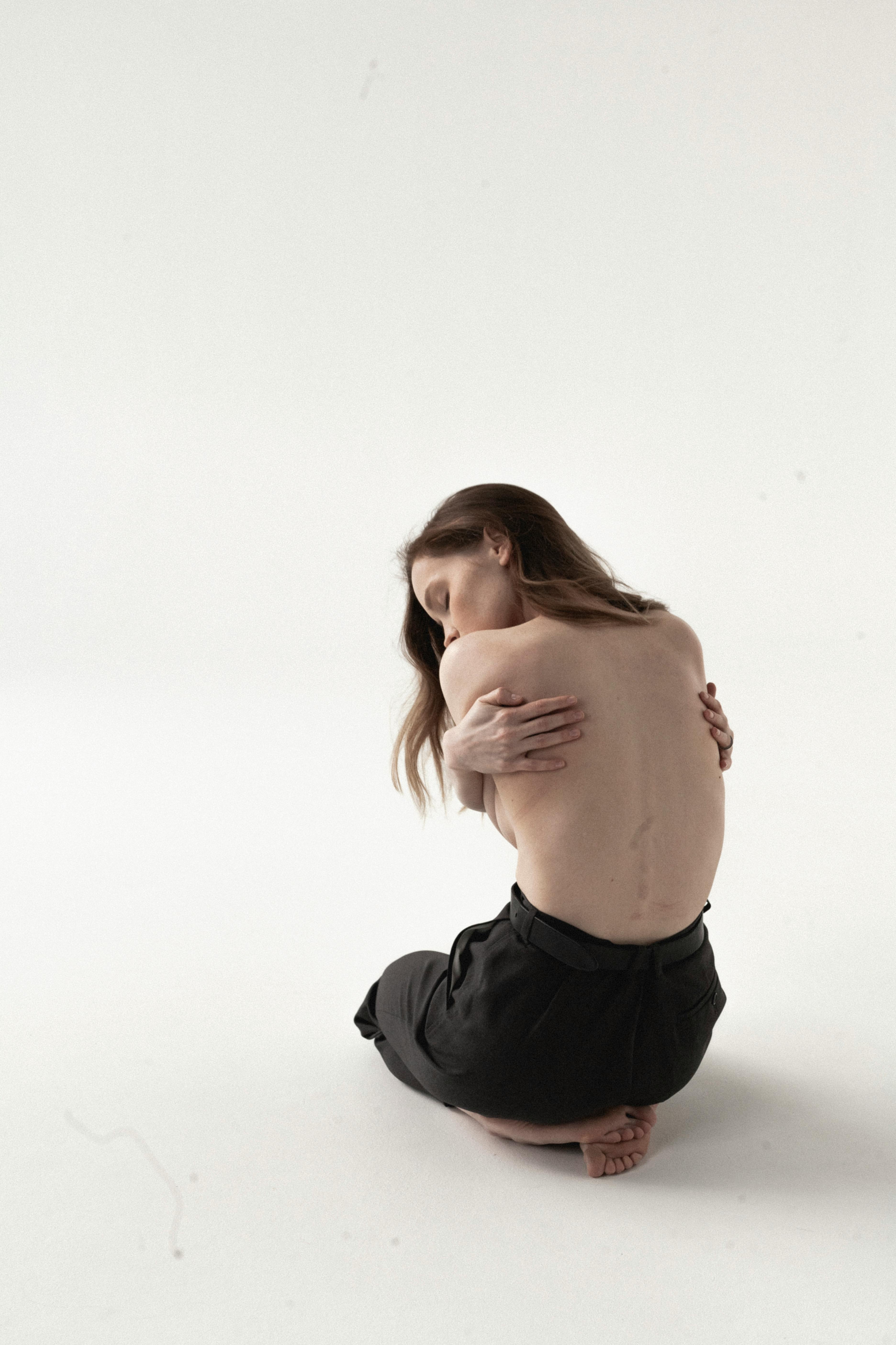 Vrouw zonder shirt zit en omarmt zichzelf, vertegenwoordigt Jungiaanse vrouwelijke archetypische beelden van kwetsbaarheid en kracht.