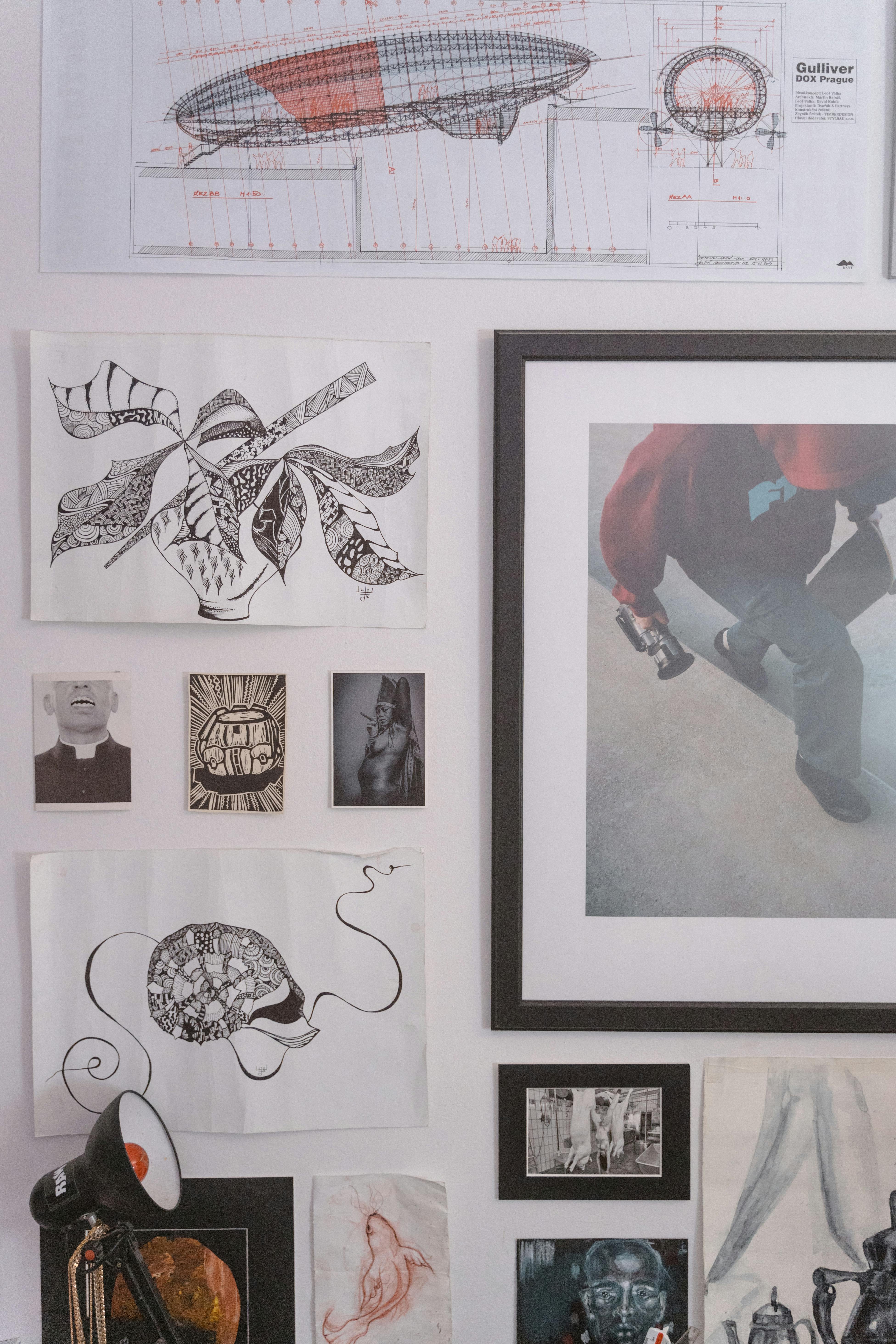 A diverse collection of artworks, including drawings, prints, and photographs, arranged on a white wall to create a gallery wall effect.
