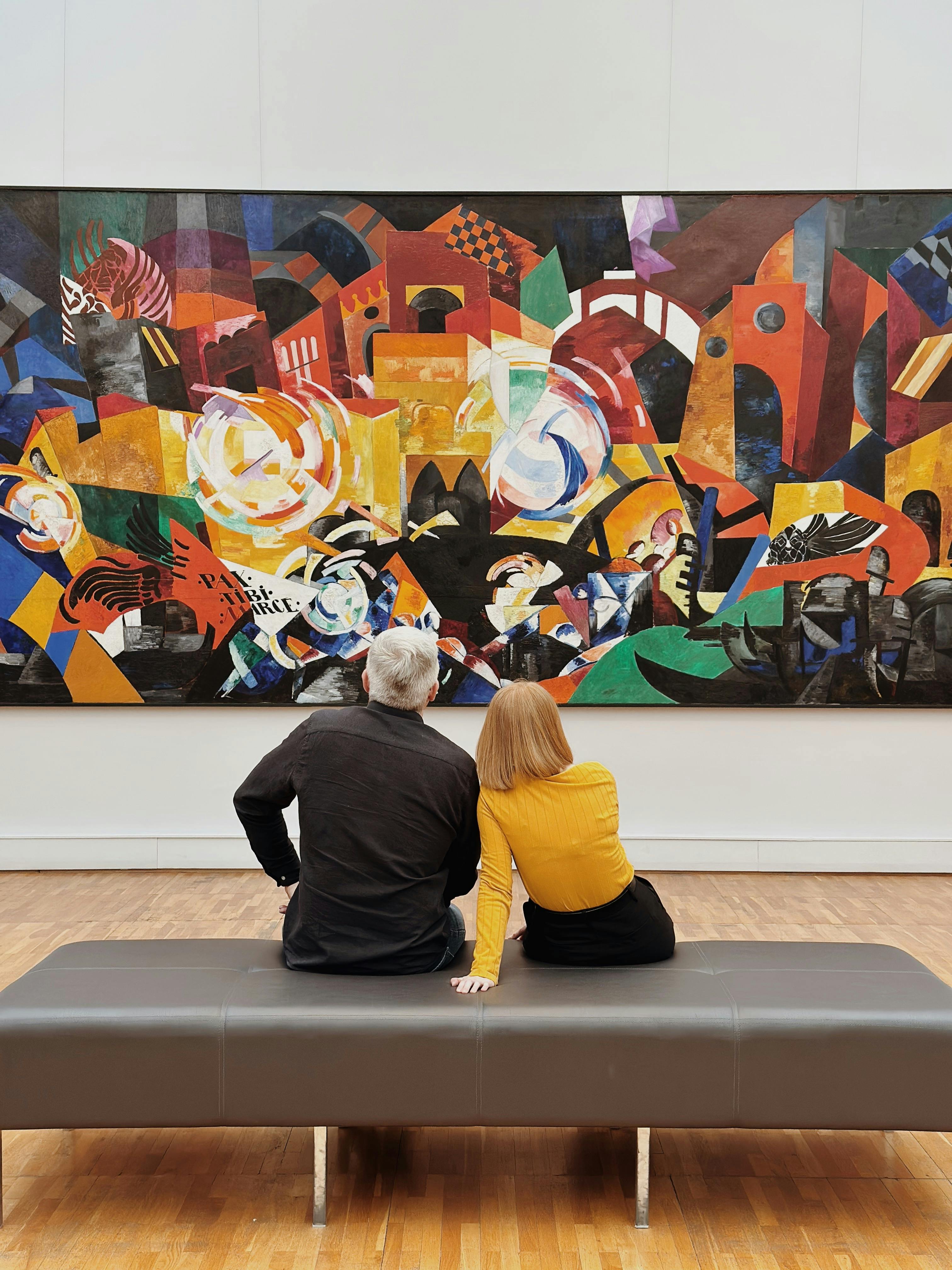 Two people examining an abstract painting in an art gallery, showcasing critical engagement with art. Man in black suit and blonde woman in yellow top observe modern artwork in a museum setting, highlighting viewer interaction with art exhibitions.
