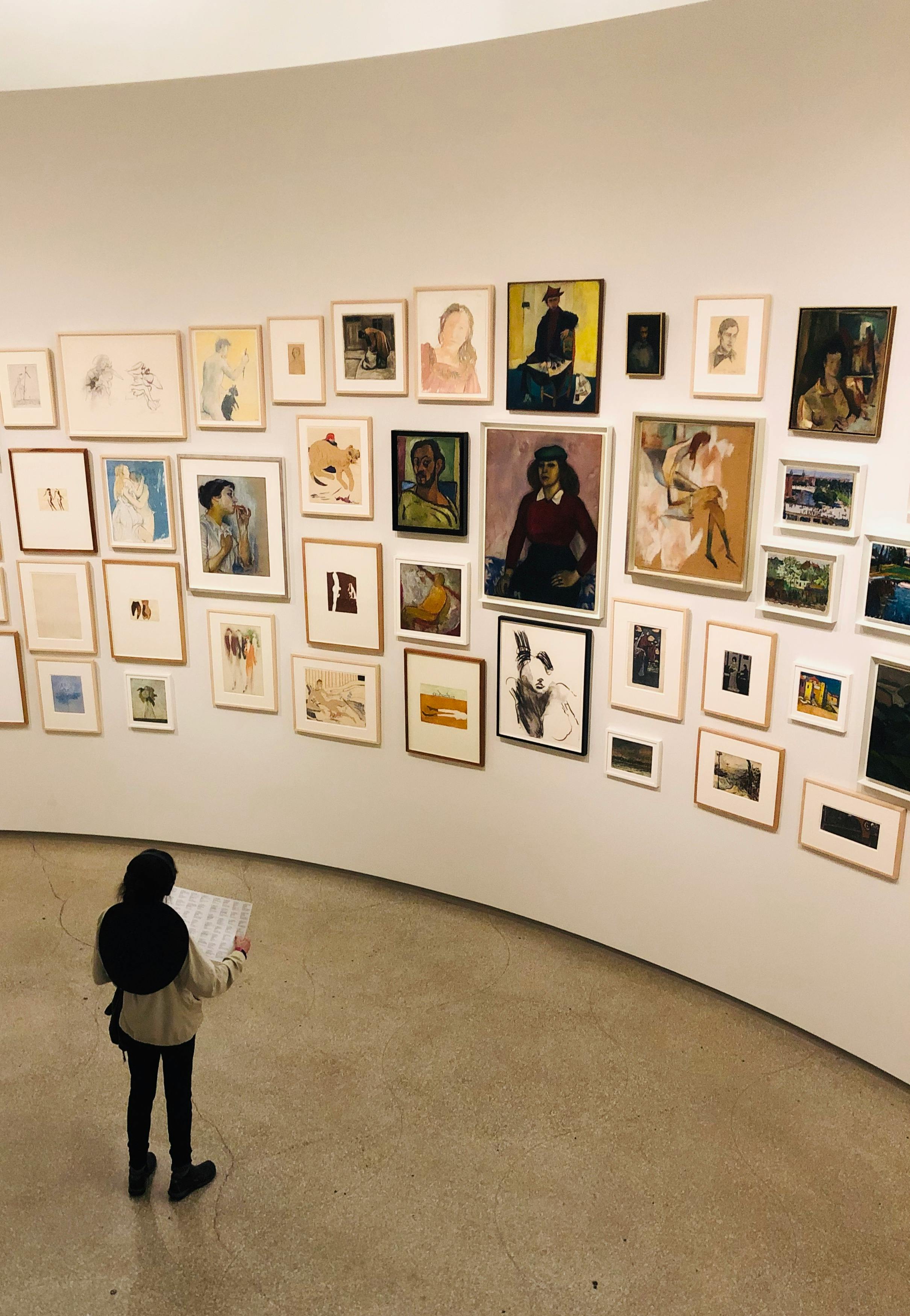A gallery visitor analyzes a comprehensive art exhibition featuring diverse framed artworks on a wall.