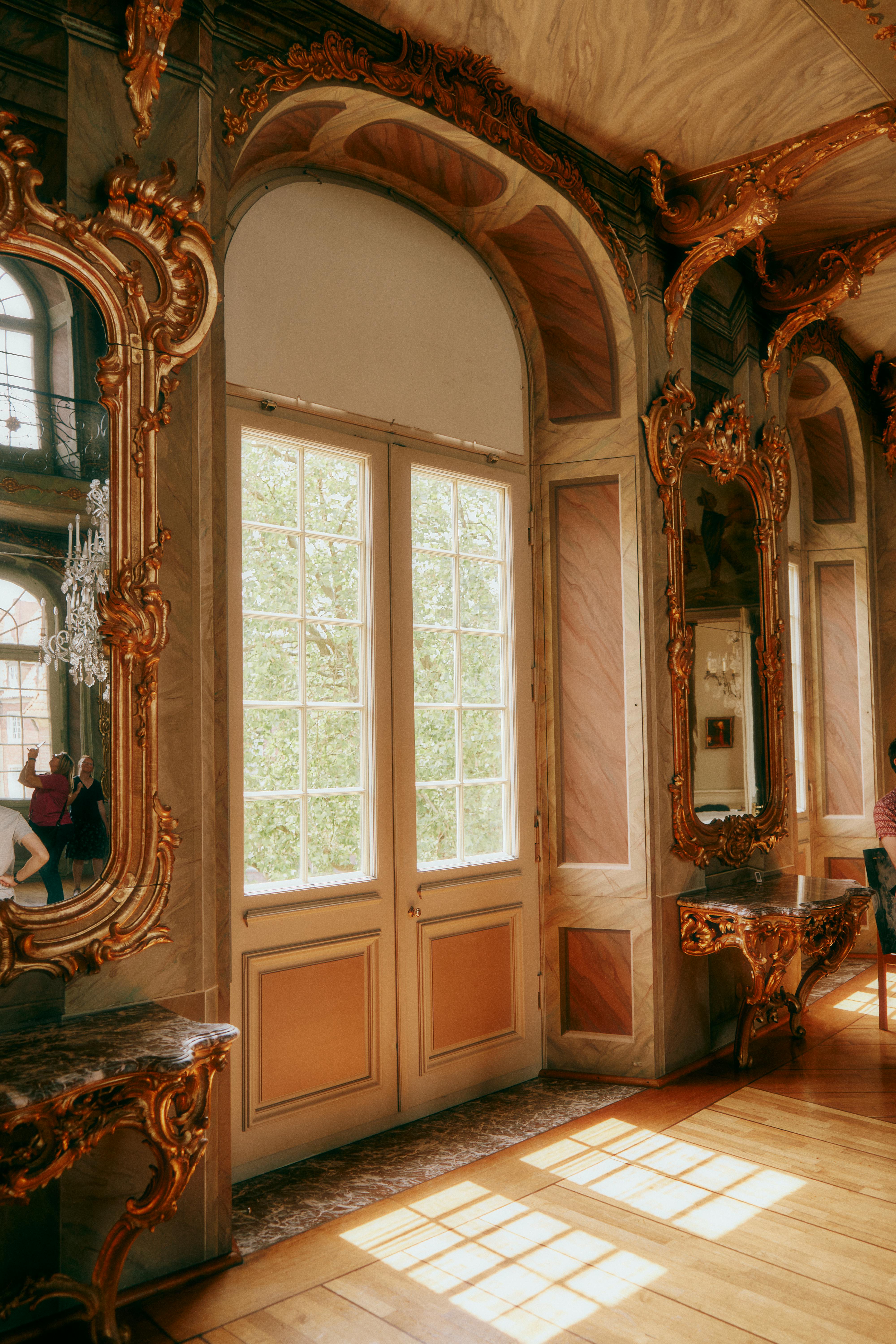 Ornate gilded mirrors and a large double door with glass panes reflecting trees and light in a room with marble walls and wooden floors.