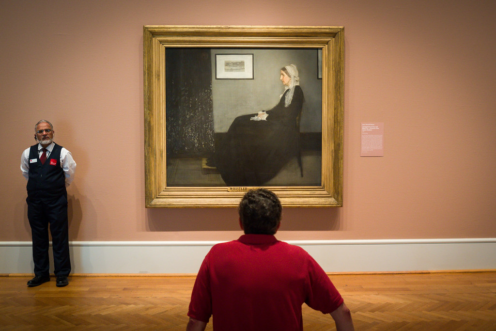 A museum guard stands to the left of James McNeill Whistler's famous painting 'Whistler's Mother', while a visitor in a red shirt views the artwork from behind.