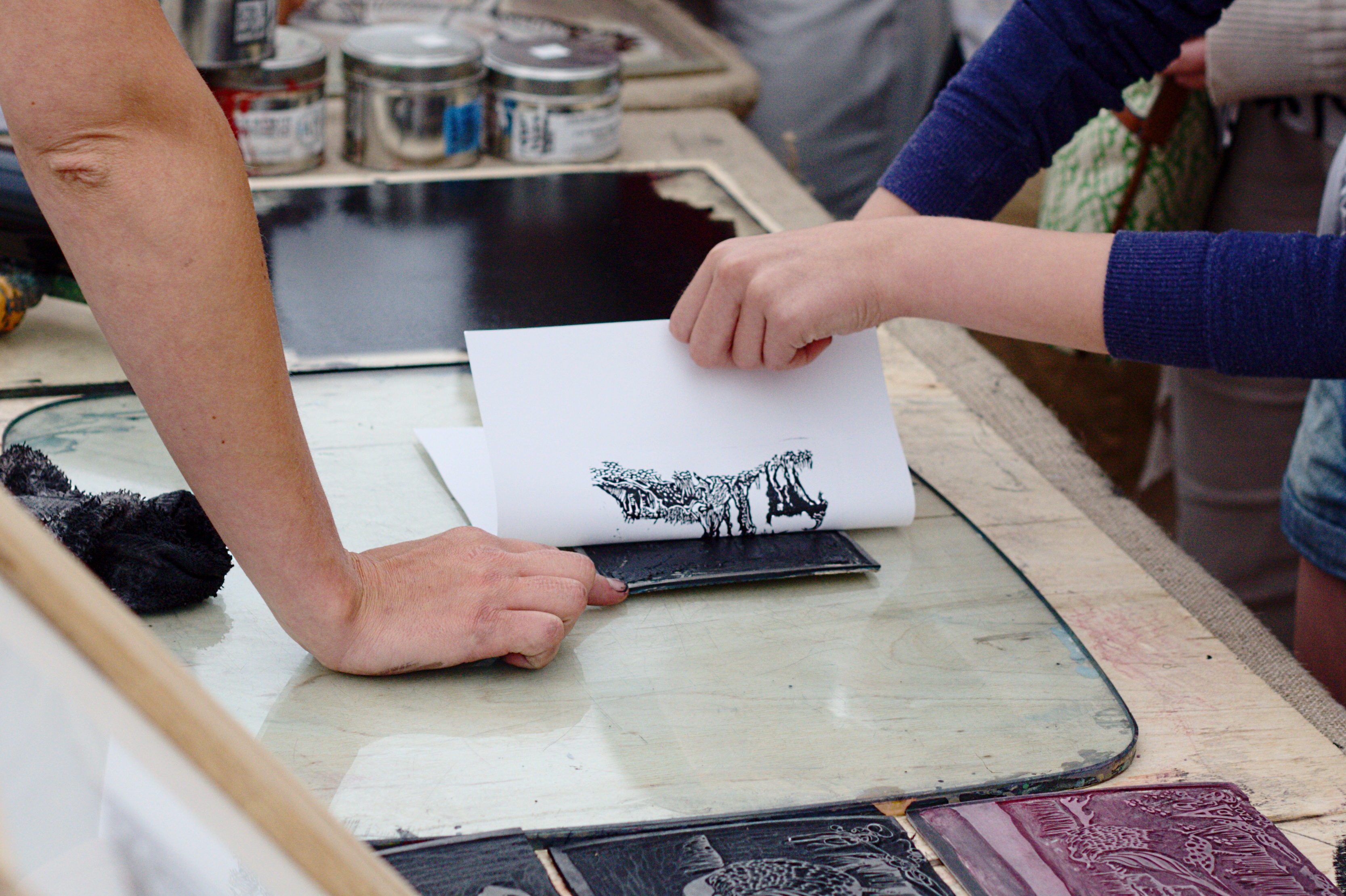 Close-up of hands performing linocut printing, showing a detailed print of a hippopotamus with its mouth open.