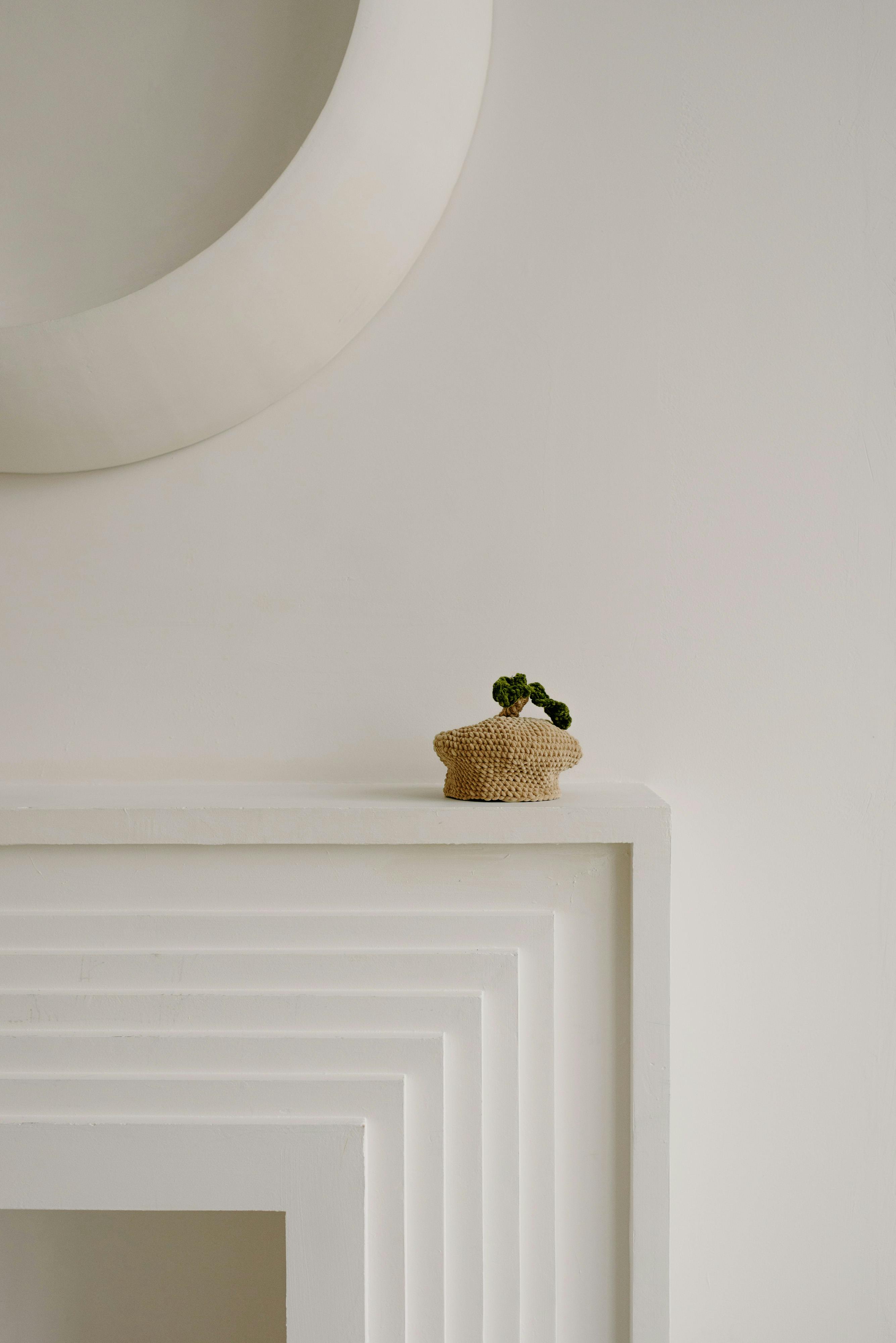 A small, crocheted decorative item resembling a plant in a pot sits on a modern, layered fireplace mantel.