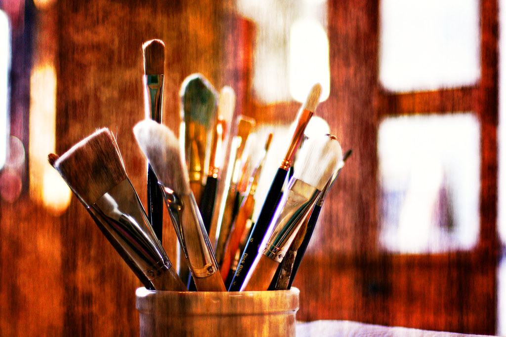 A collection of various paint brushes, some with thick bristles and others with finer tips, arranged in a wooden holder, suggesting essential tools for digital painting.