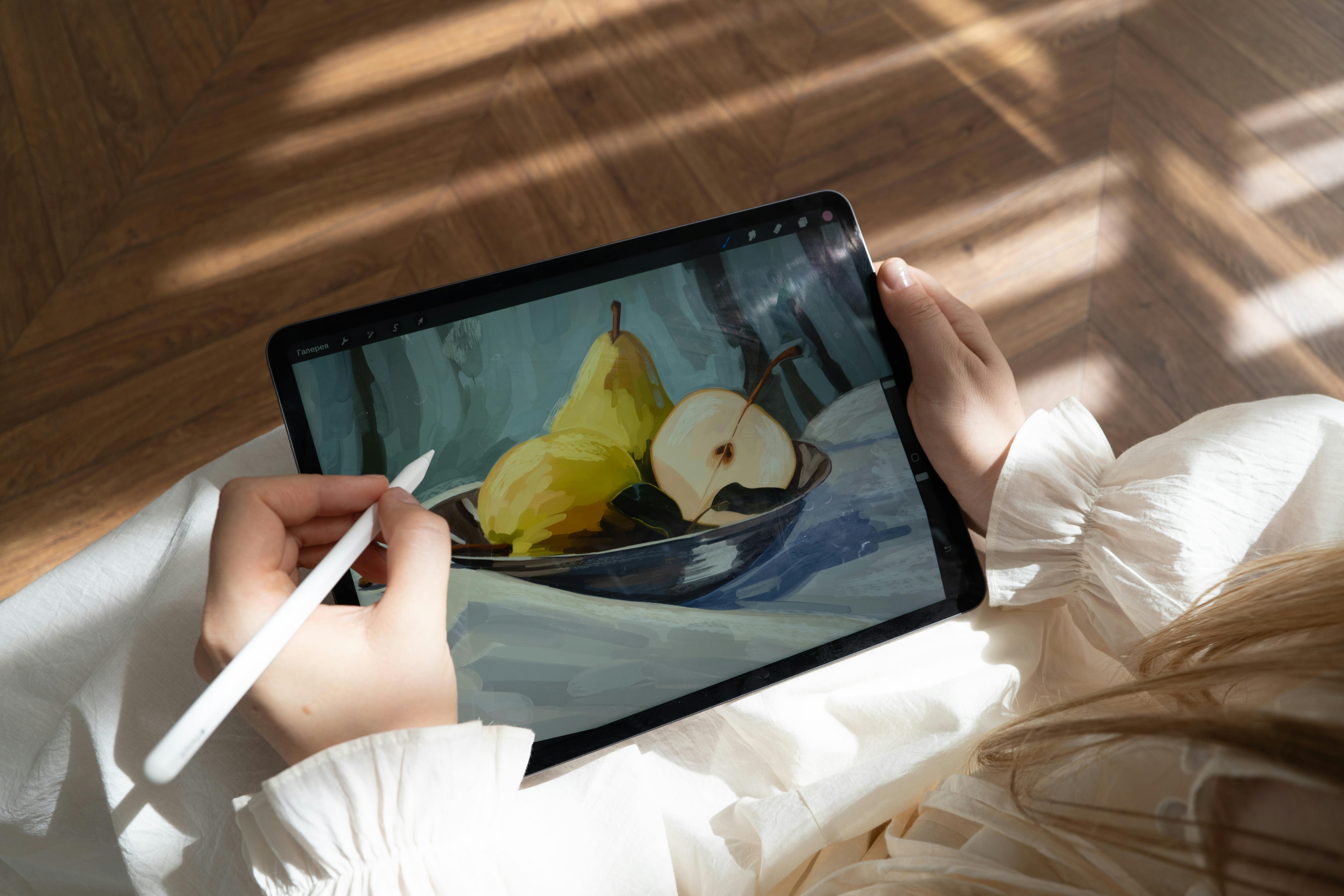 A girl with blonde hair wearing a white ruffled shirt is drawing on a tablet with a stylus. The tablet displays a digital painting of pears in a bowl, with sunlight casting shadows on the wooden floor.