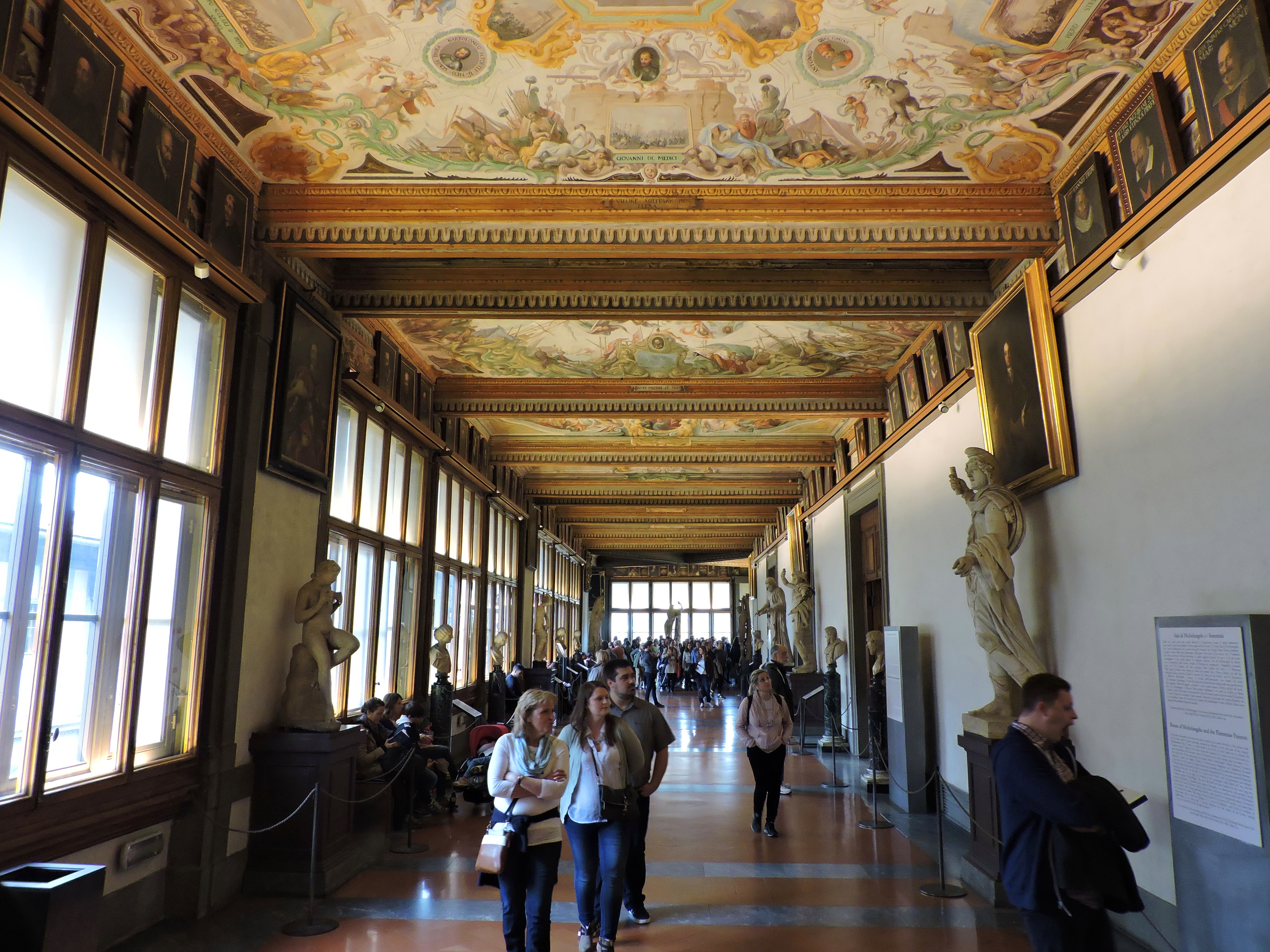 Interior view of the Uffizi Gallery in Florence, showcasing a long hallway adorned with paintings and sculptures, with visitors walking through.