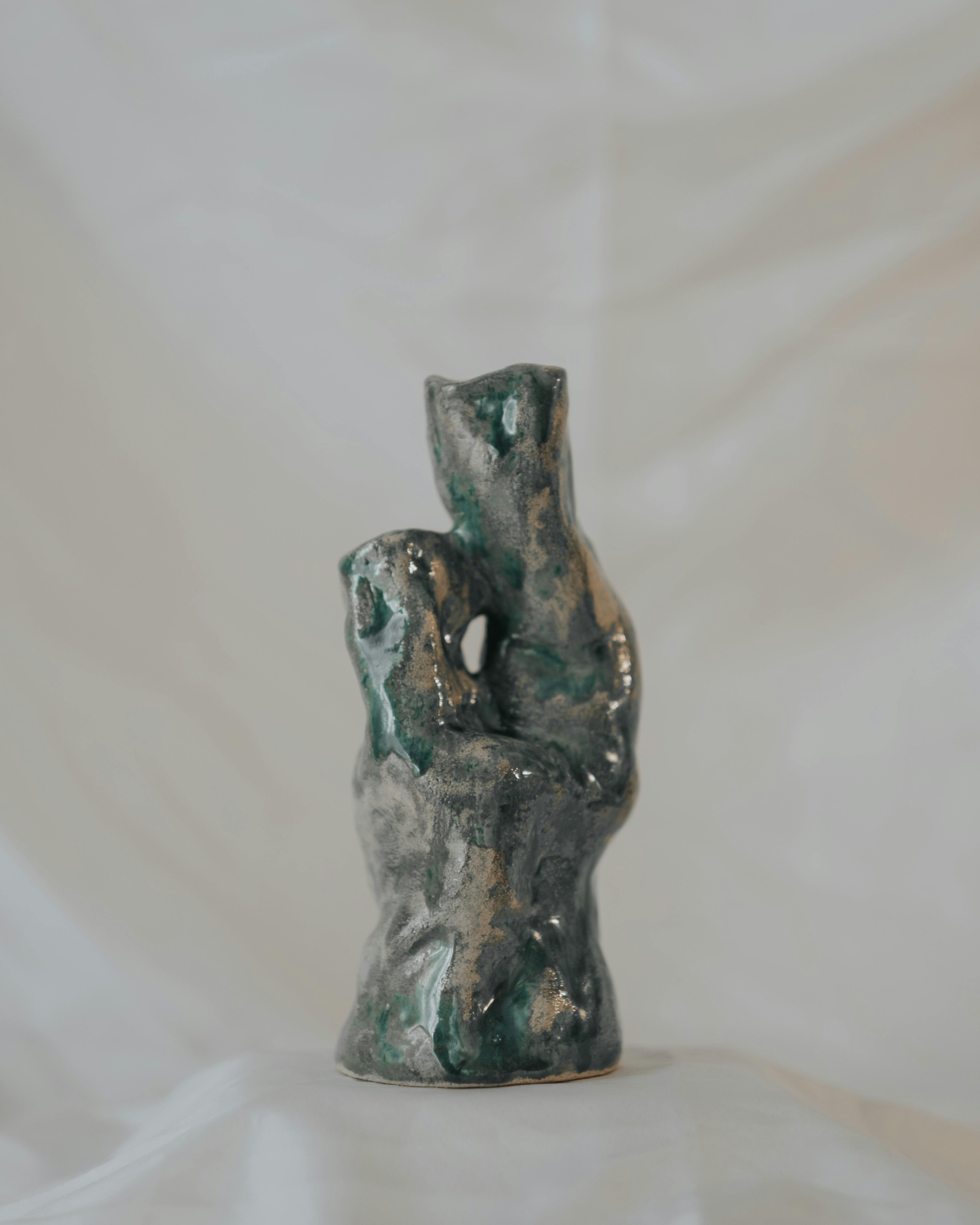 Abstract keramisch sculptuur met groen en grijs glazuur, getoond in zachte verlichting tegen een witte achtergrond.