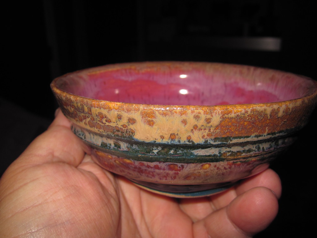 Een hand houdt een kleine, kleurrijke keramische kom vast met een roze binnenkant en een getextureerd, veelkleurig glazuur aan de buitenkant.