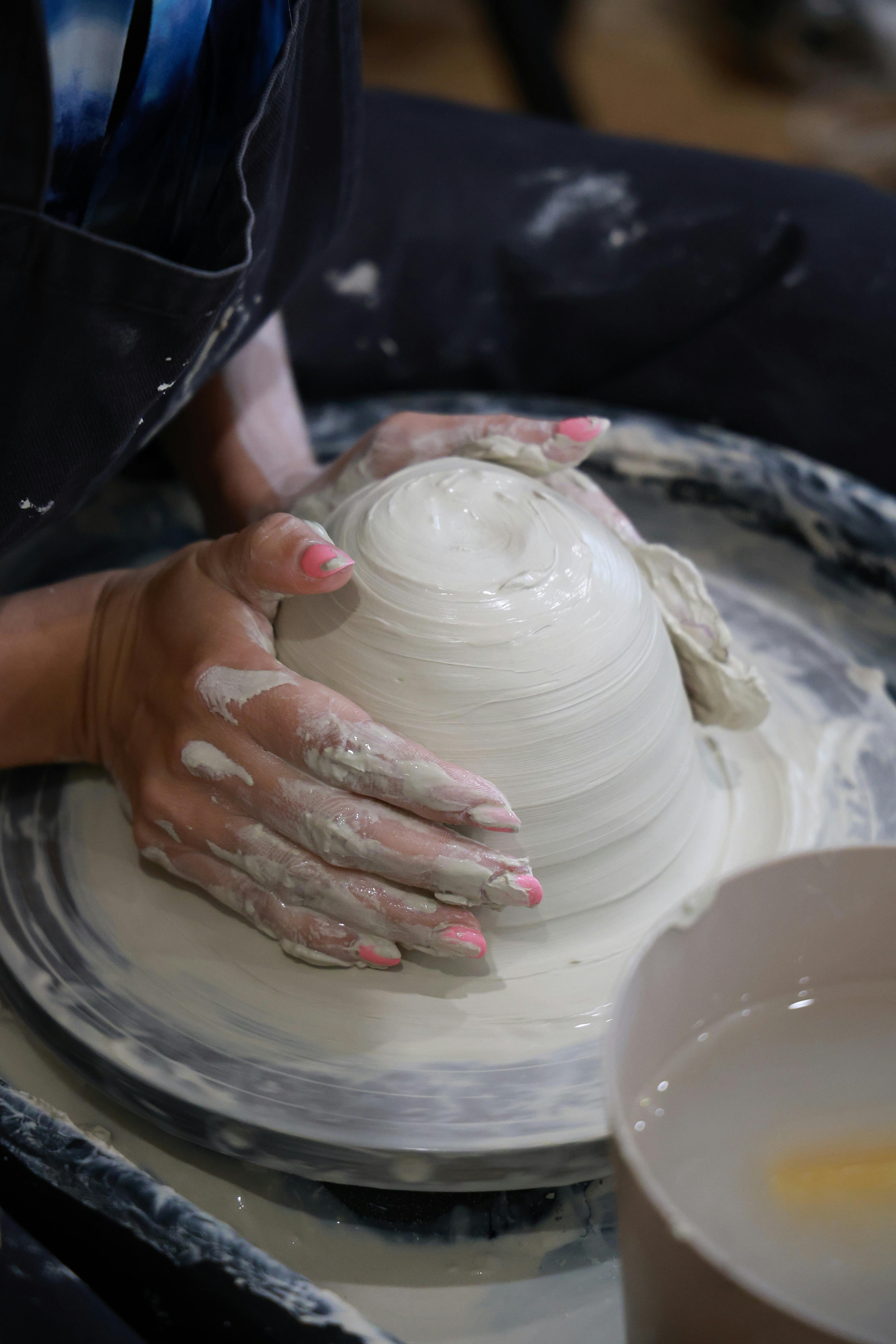 De handen van een persoon met roze gelakte nagels vormen een natte kleikom op een pottenbakkerswiel, met een kom water ernaast.