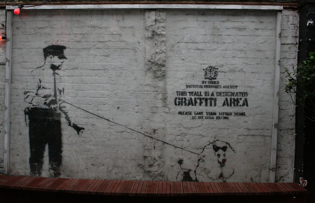 Banksy's 'Designated Graffiti Area' artwork featuring a police officer walking a poodle with a red nose, painted on a white brick wall with official-looking text.