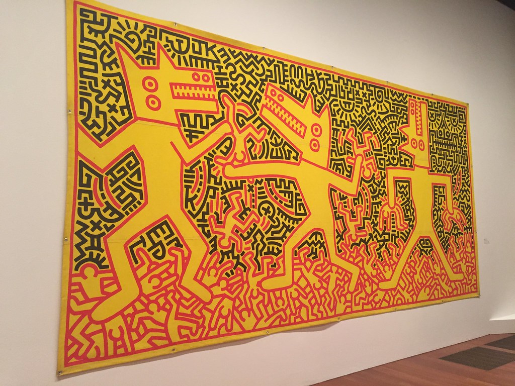 Large Keith Haring artwork featuring three yellow dog-like figures with black outlines and red details, set against a background of black and red abstract patterns, displayed on a white wall in a museum.