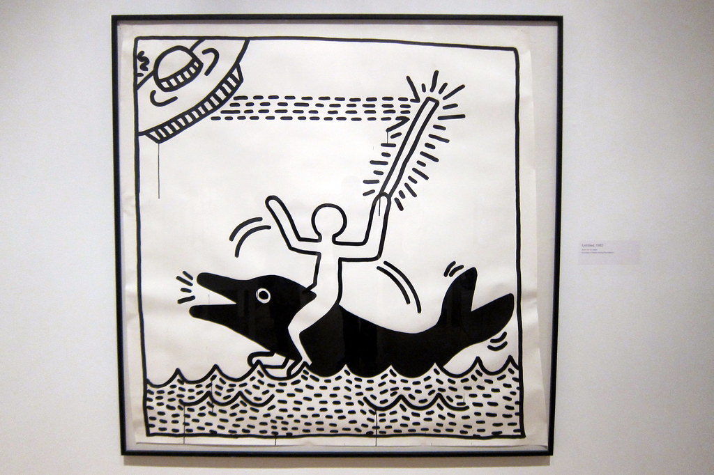 Black and white Keith Haring artwork featuring a stick figure riding a dolphin in water, holding a glowing stick, with a large hat-like object above. The piece showcases Haring's iconic bold line style.