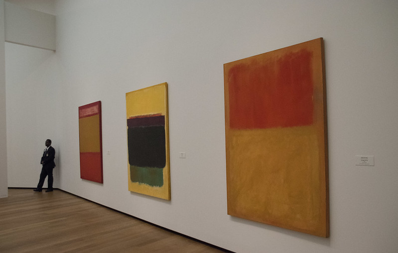 View of three large-scale color field paintings by Mark Rothko displayed on a white wall in the Fondation Beyeler museum, with a visitor standing to the left.