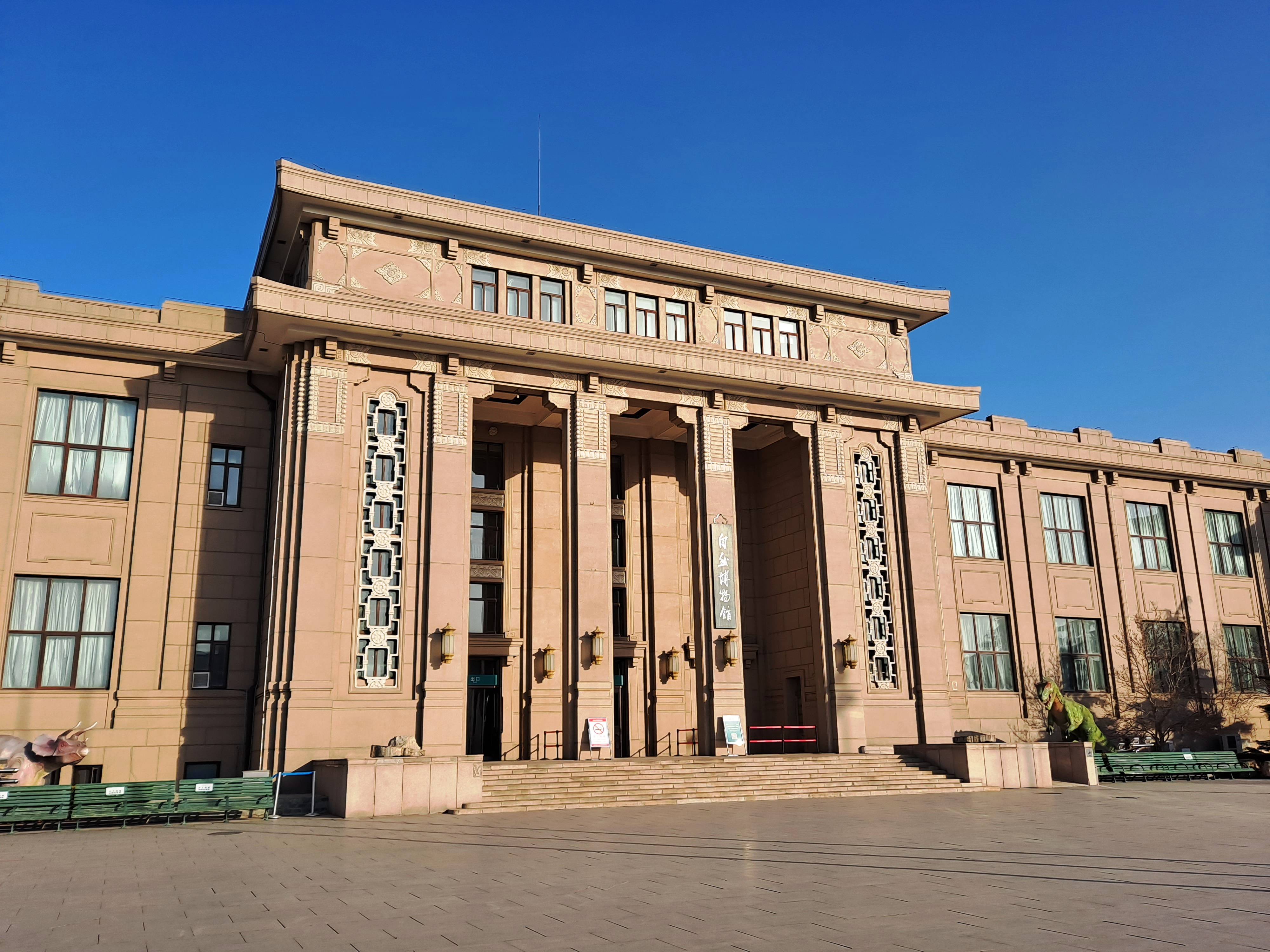 Front view of the Beijing Museum of Natural History, a grand beige building with classical architecture and columns, under a clear blue sky, with dinosaur statues on the plaza.