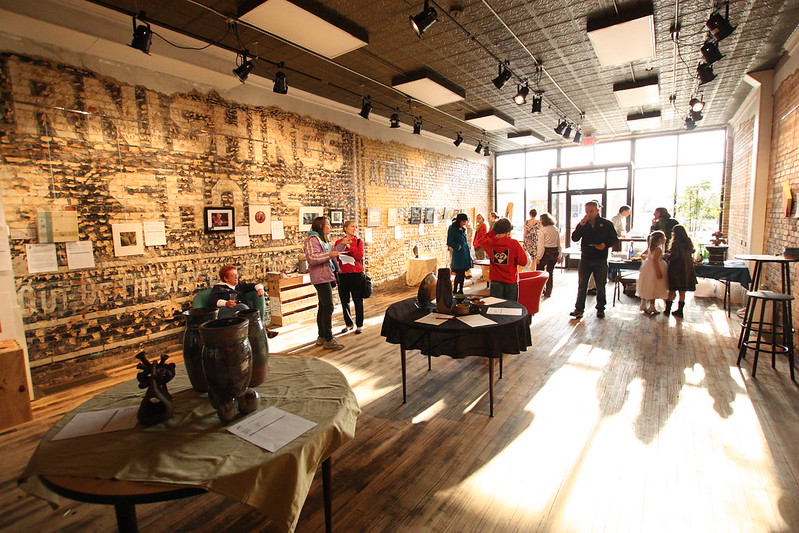 Interior view of a bustling local art gallery during an opening reception. People are mingling, observing the displayed artworks on the brick walls and tables, under track lighting and natural light streaming through the front windows.