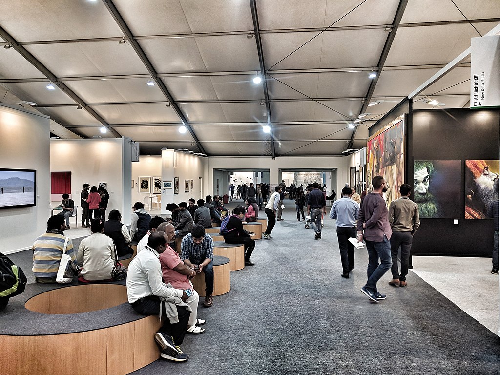 Interieurzicht van een drukke kunstbeurs met veel mensen die verschillende kunstwerken bekijken die langs de muren en in stands worden tentoongesteld.